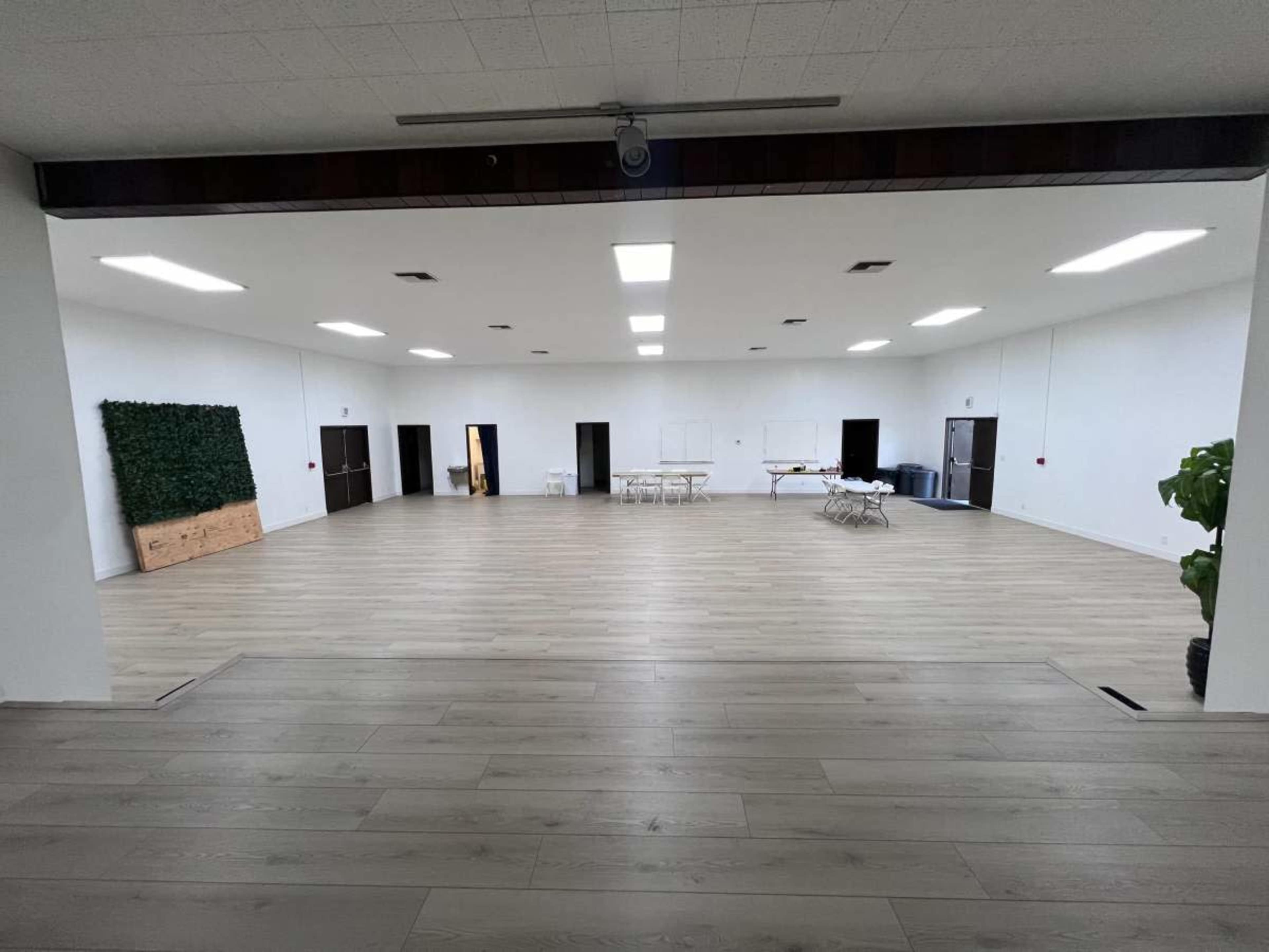 The image shows a large, empty room with light-colored wooden flooring, white walls, and several tables and chairs set up in the center.