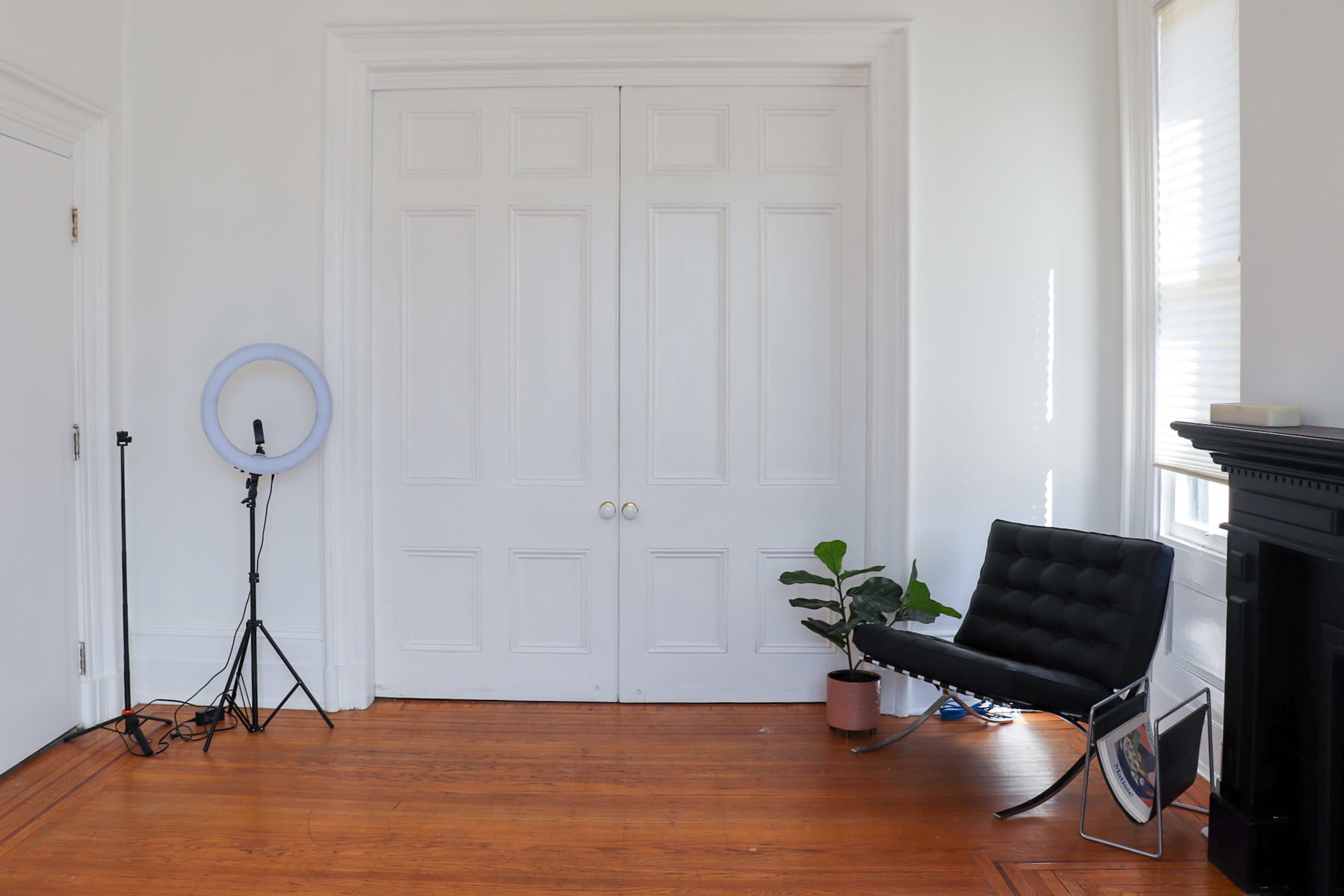 The image shows a minimalist room with white walls, a black chair, a ring light on a stand, a potted plant, and large double doors.
