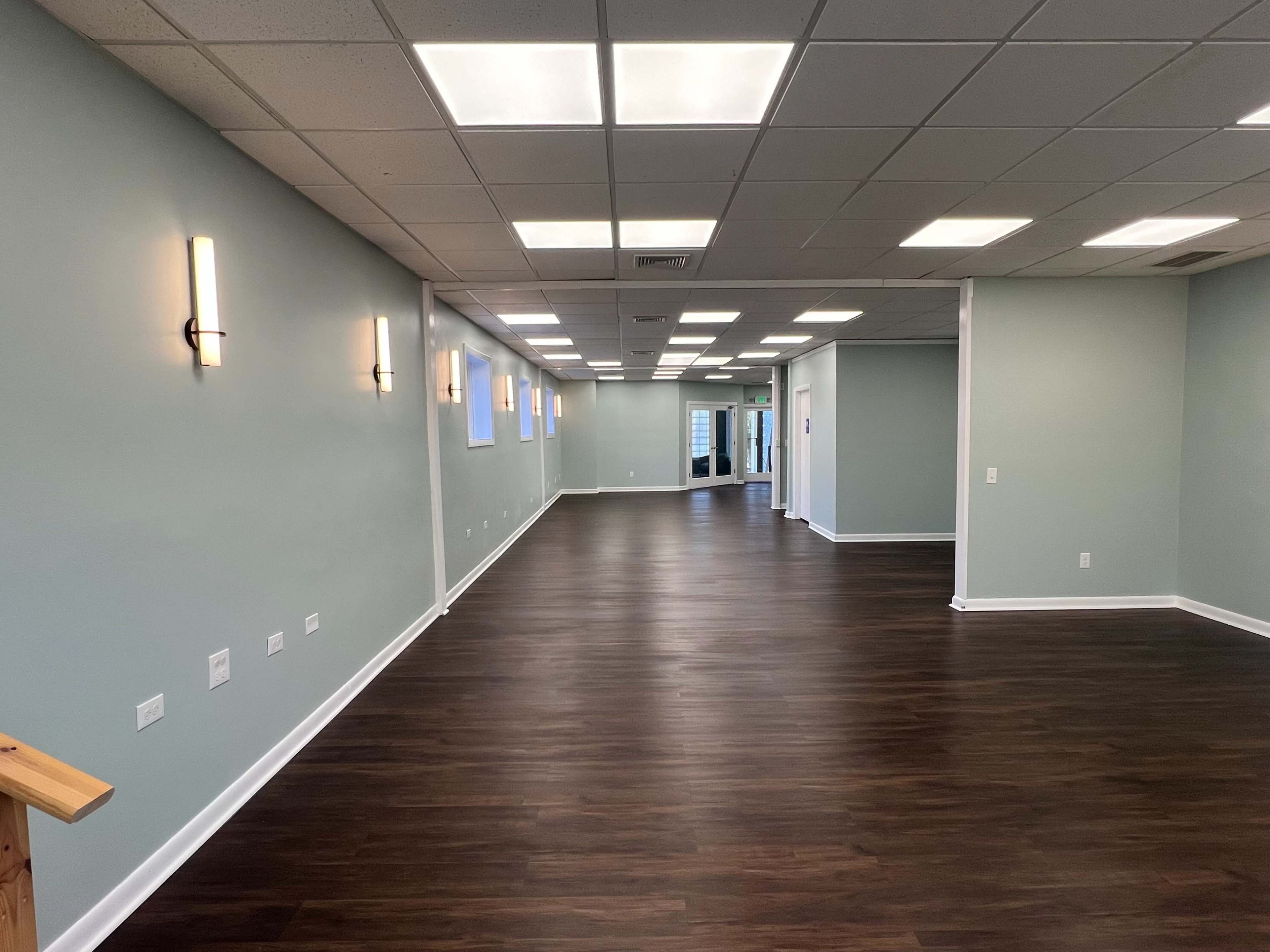 The image shows a large, empty room with a wooden floor, light green walls, and overhead fluorescent lighting.