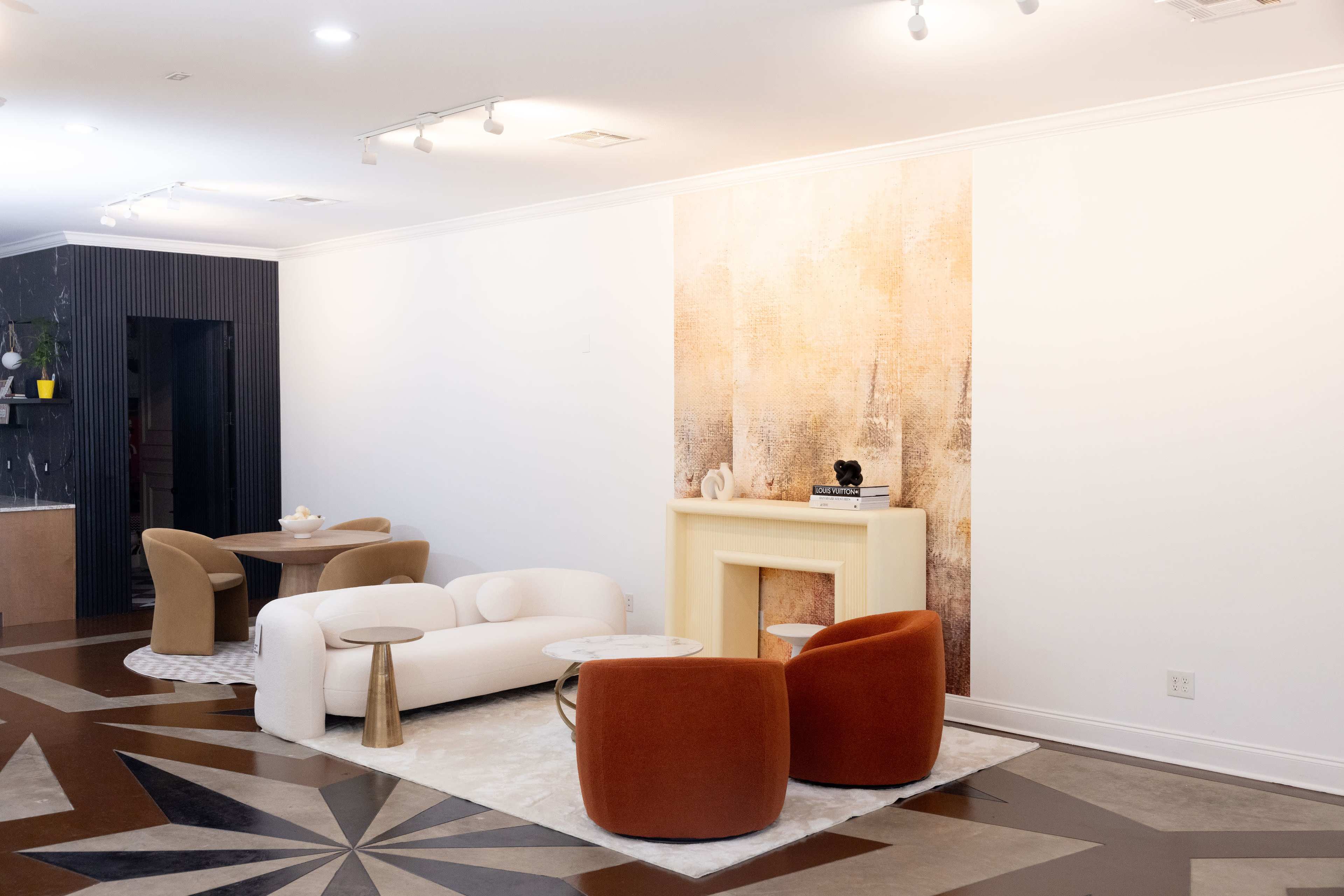 The image features a modern interior space with a white sofa, two brown armchairs, a small round table, and a decorative fireplace against a textured wall.