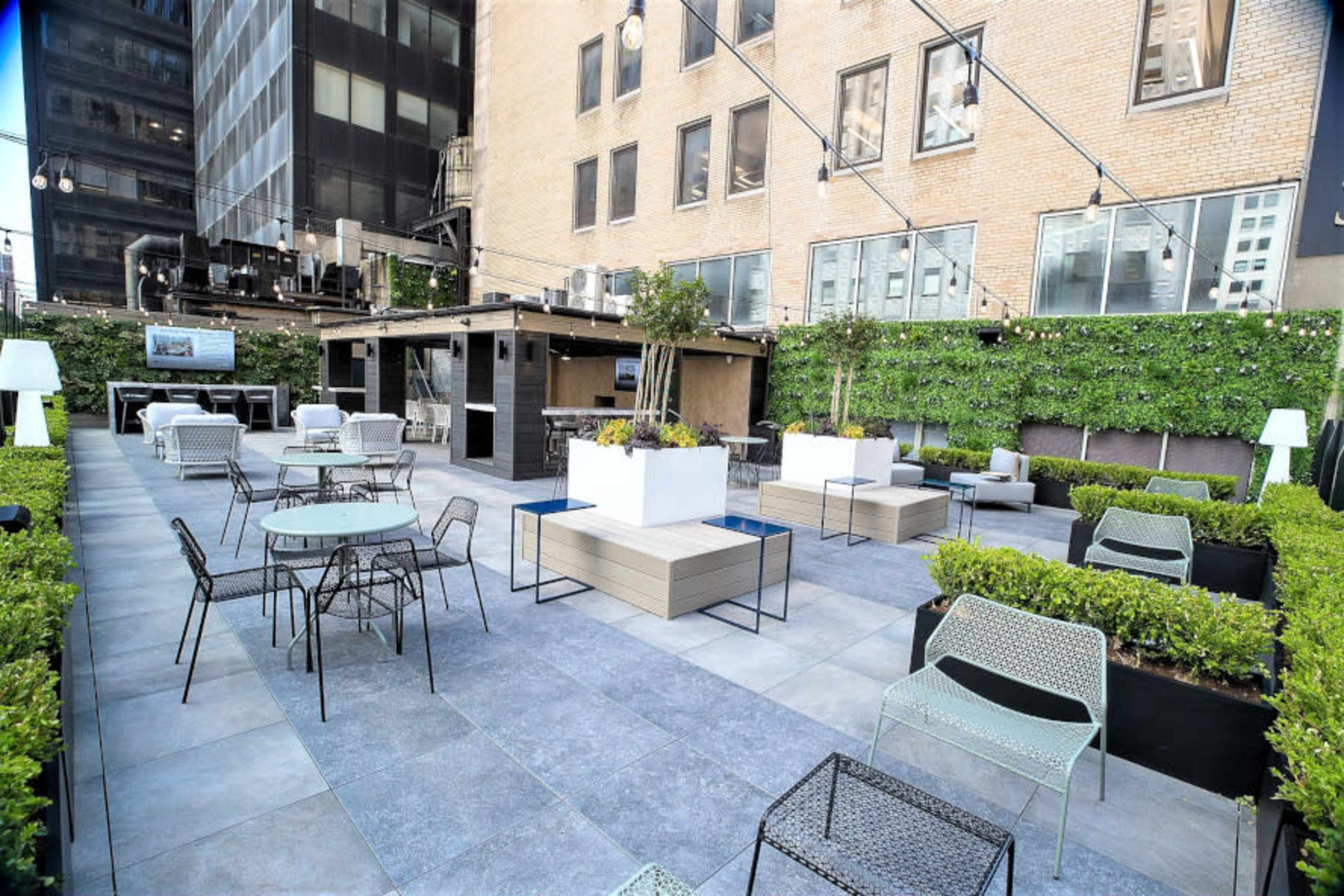 The image shows a modern outdoor patio area featuring seating arrangements, planters, and a bar, surrounded by tall buildings.