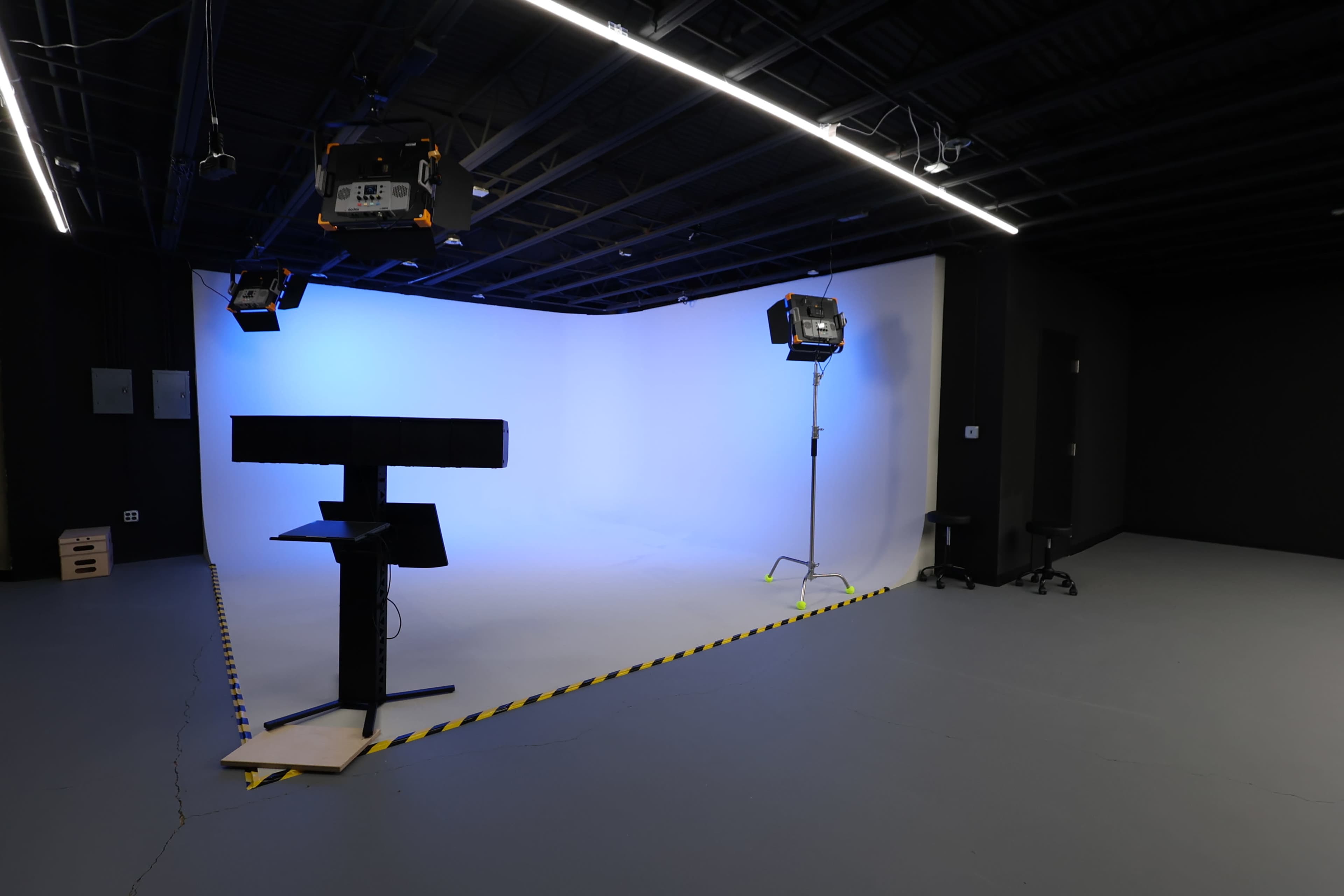 The image shows a photography or filming studio with a seamless light blue backdrop, lighting equipment overhead, and minimal furniture in a dimly lit space.