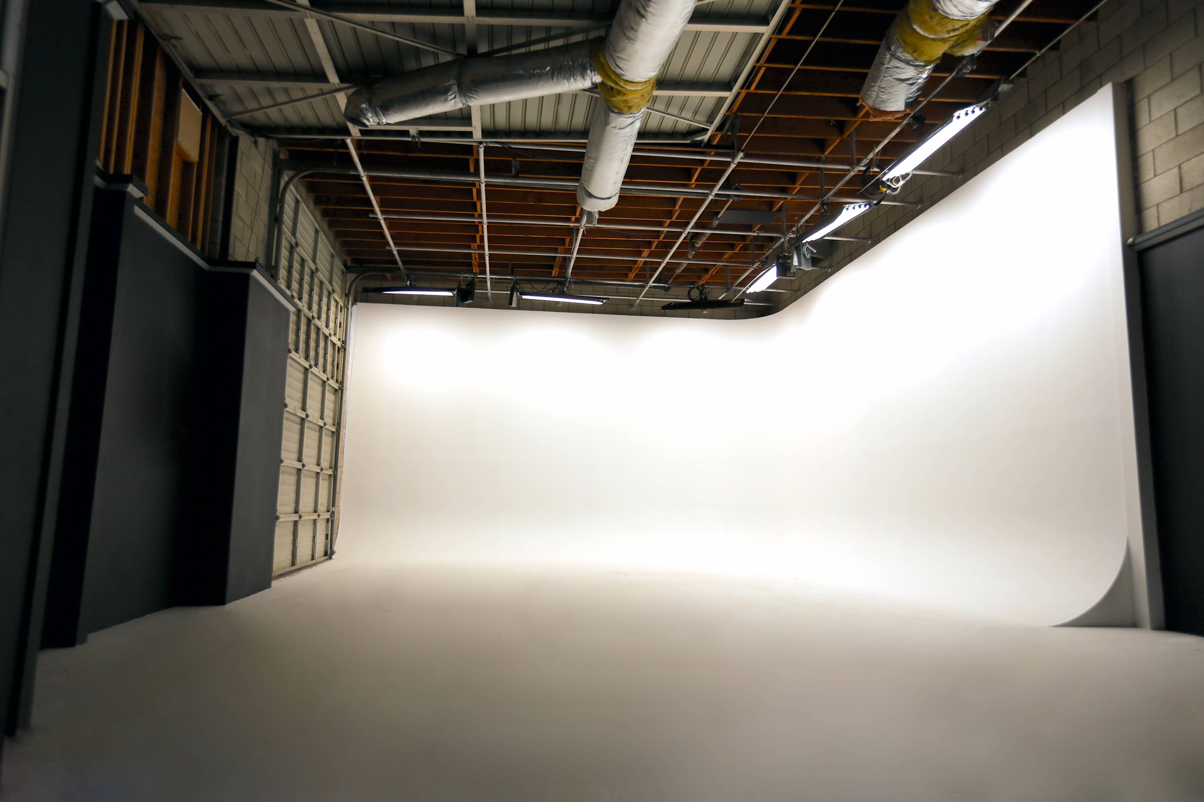 A spacious, empty photography studio with a large seamless white backdrop and exposed ceiling features.