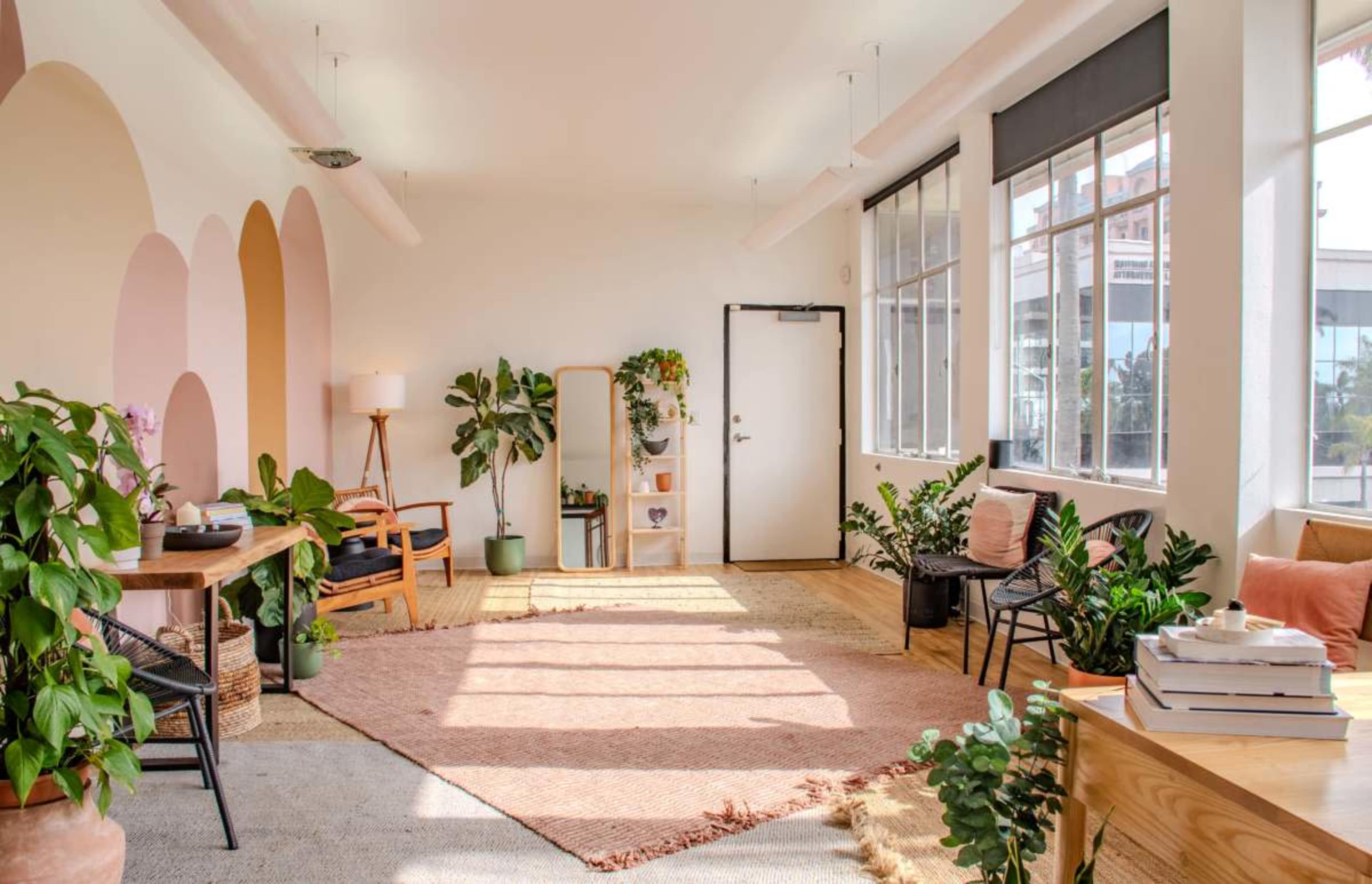 The room features a spacious layout with large windows, plants in various corners, a potted tree, and a neutral-colored rug on the floor.