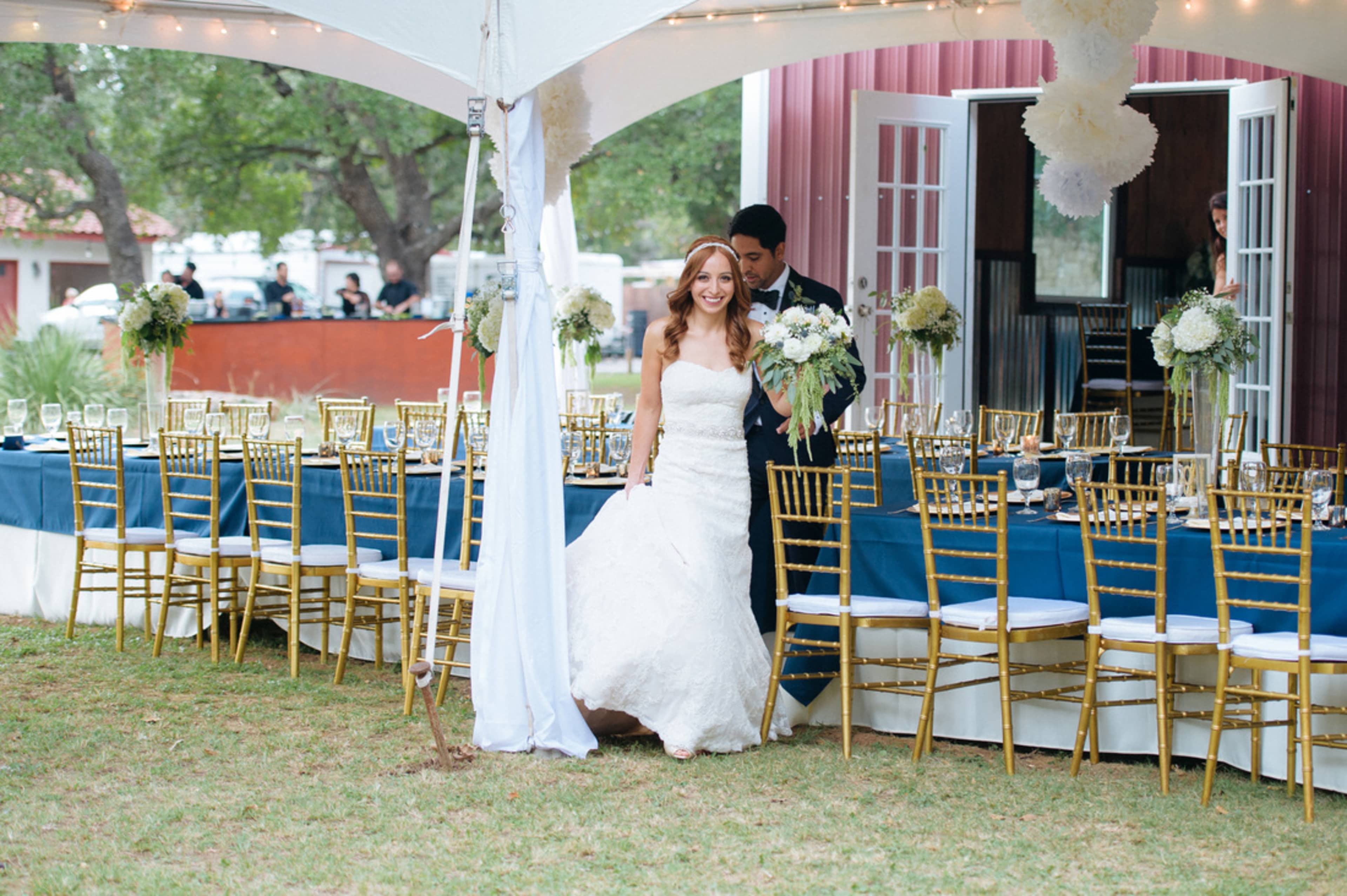 A bride in a white dress walks into a tented reception area with a groom, surrounded by elegantly arranged tables and golden chairs.