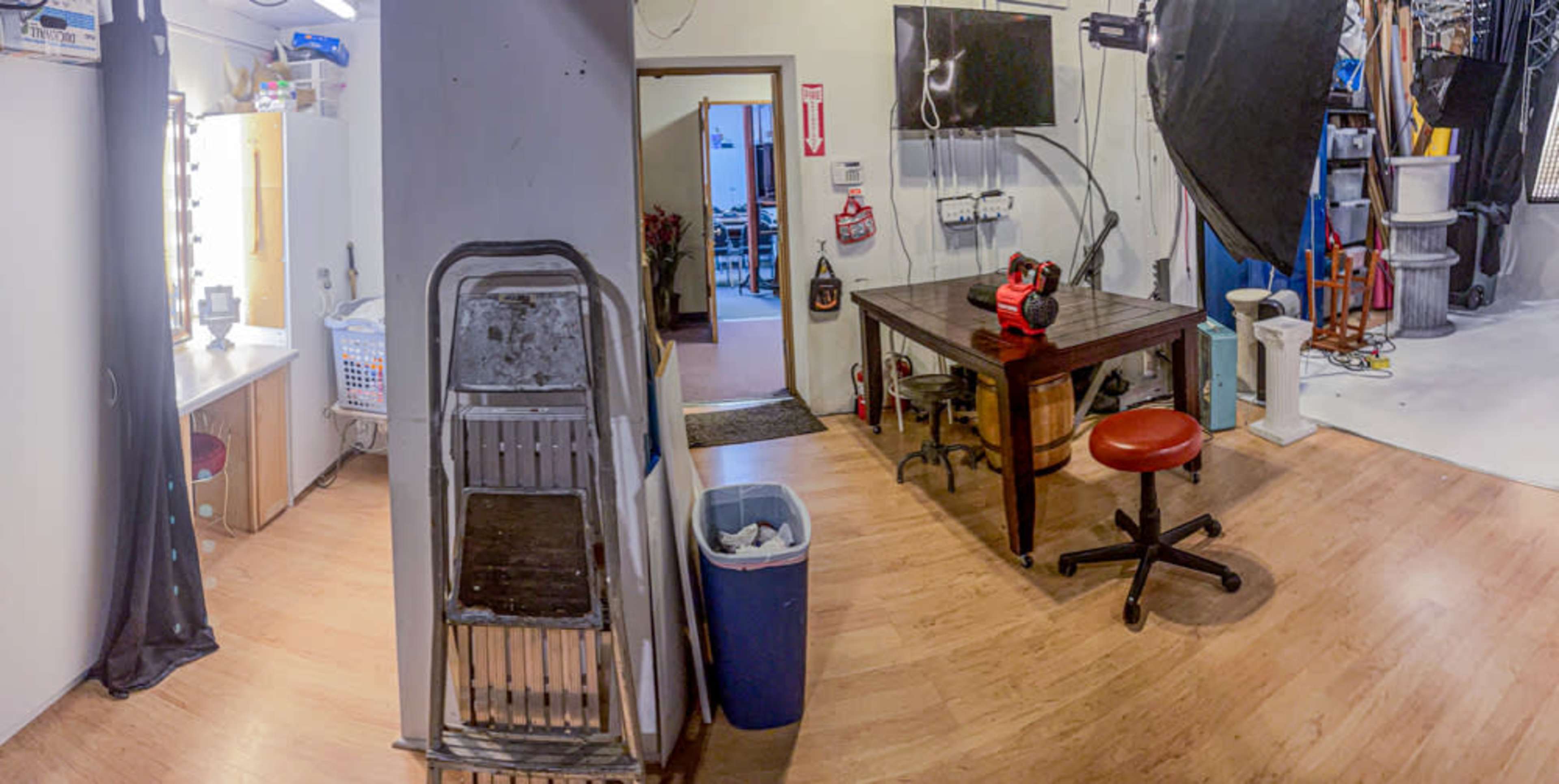 The image shows a studio space featuring a dressing area with mirrors, a desk with equipment in the center, and a photography backdrop setup in the background.