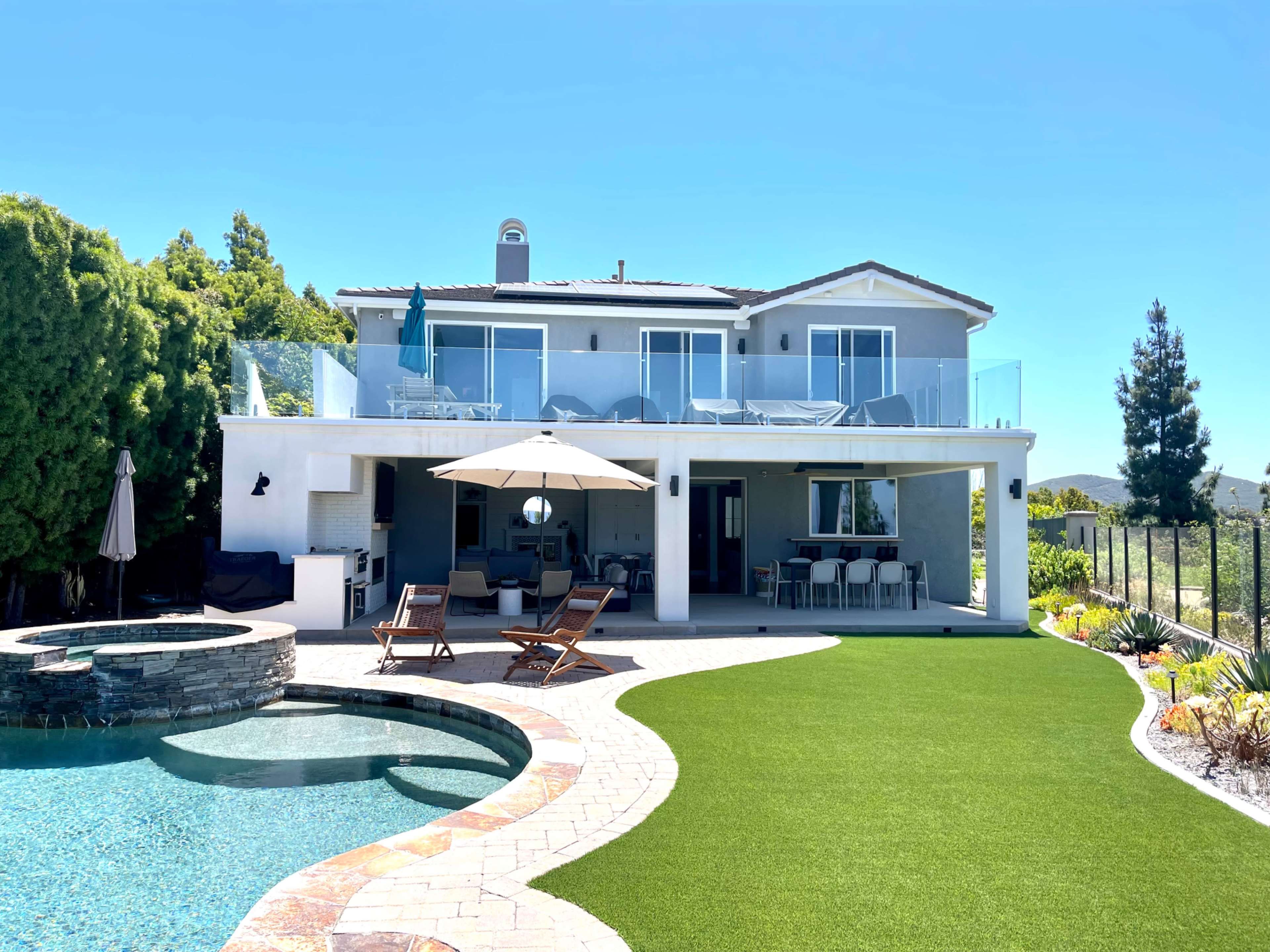 A two-story house with a patio overlooking a pool, surrounded by landscaped greenery.