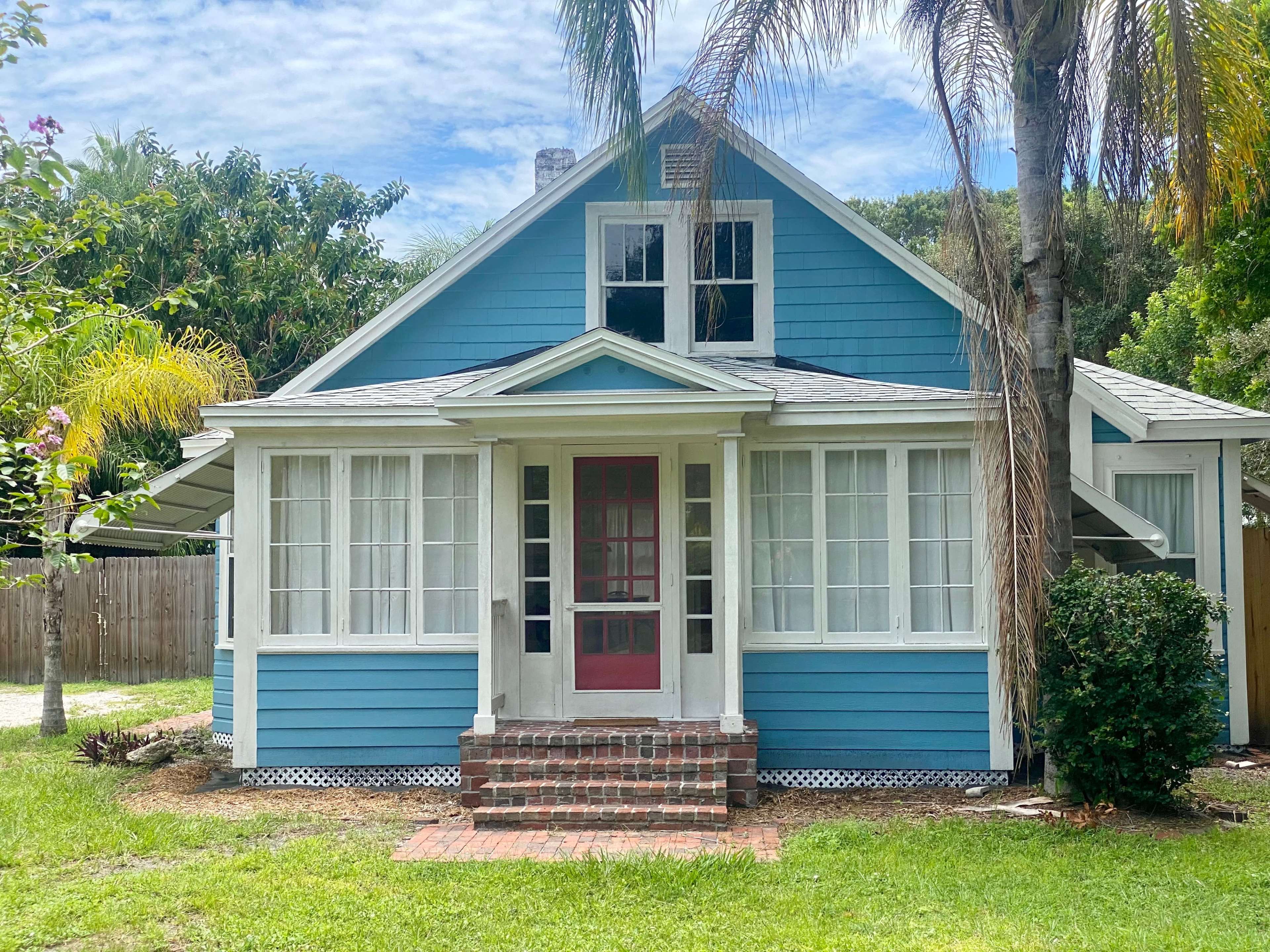 A blue house with white trim and a red front door, featuring a wide porch and surrounded by green grass and palm trees.