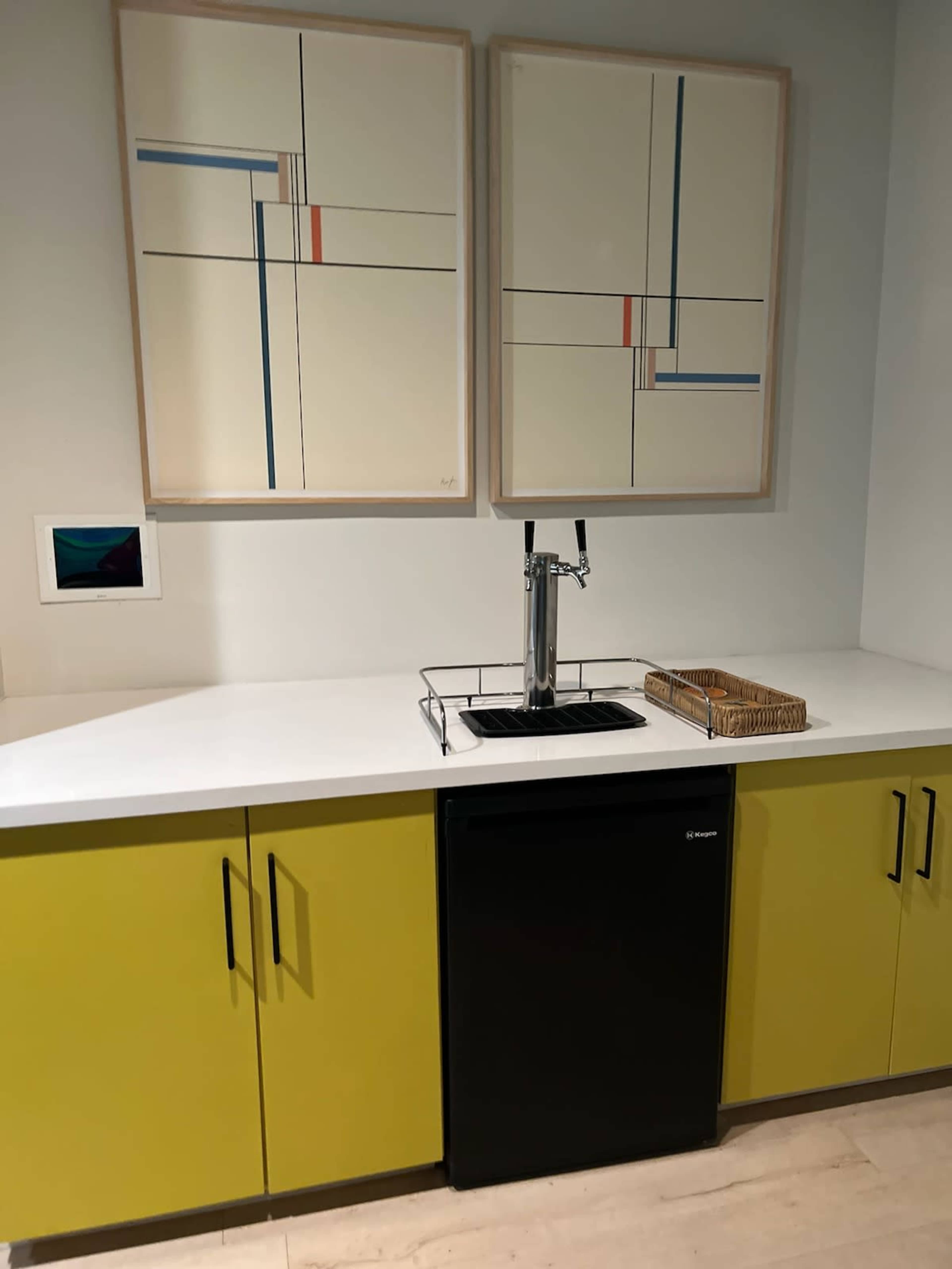 A kitchen area with a black mini refrigerator, a white countertop, a wall-mounted faucet on a tray, and two abstract framed artworks above.