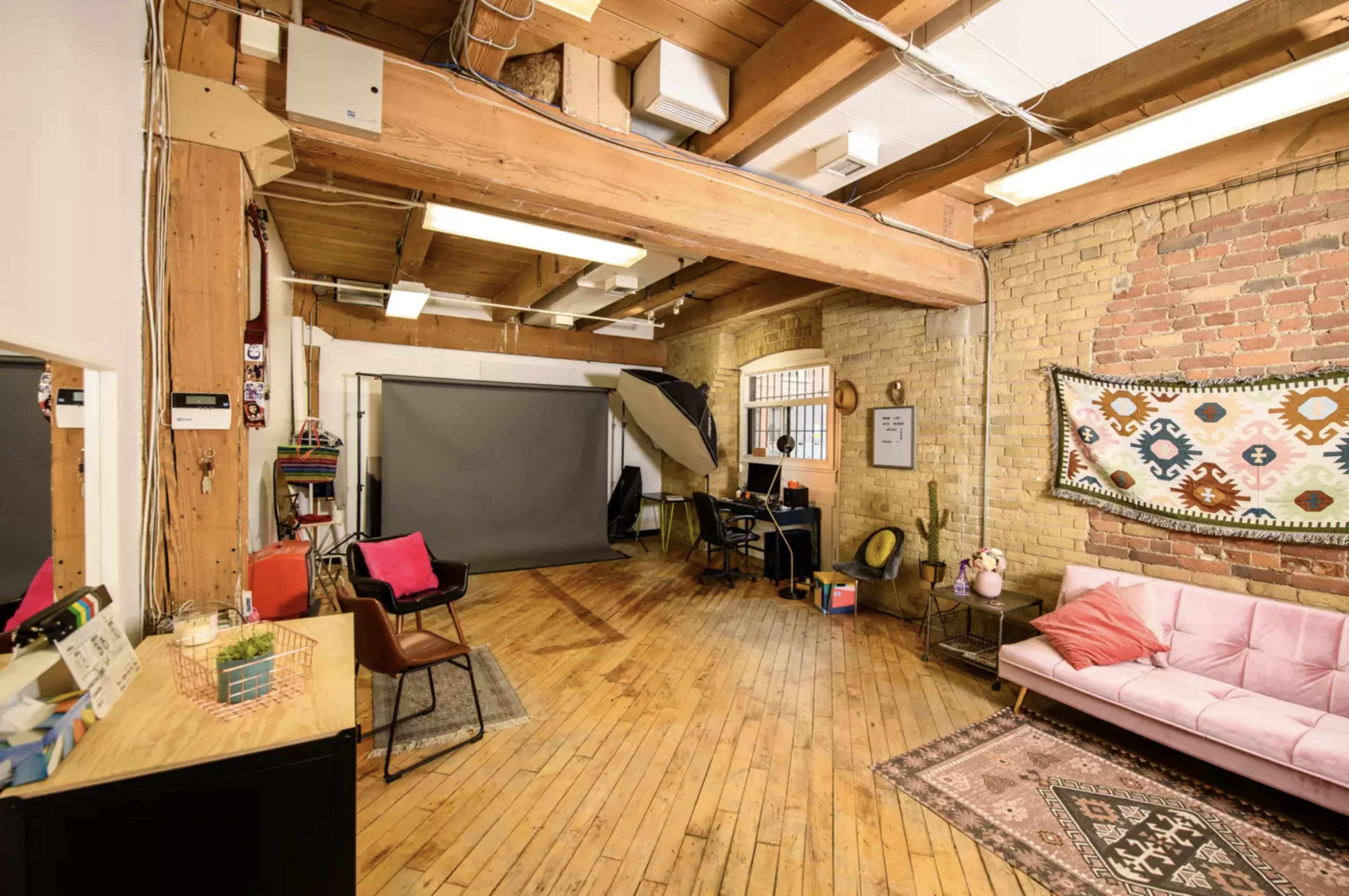 A spacious studio with wooden beams, a large gray backdrop, a pink sofa, and a desk with a chair.