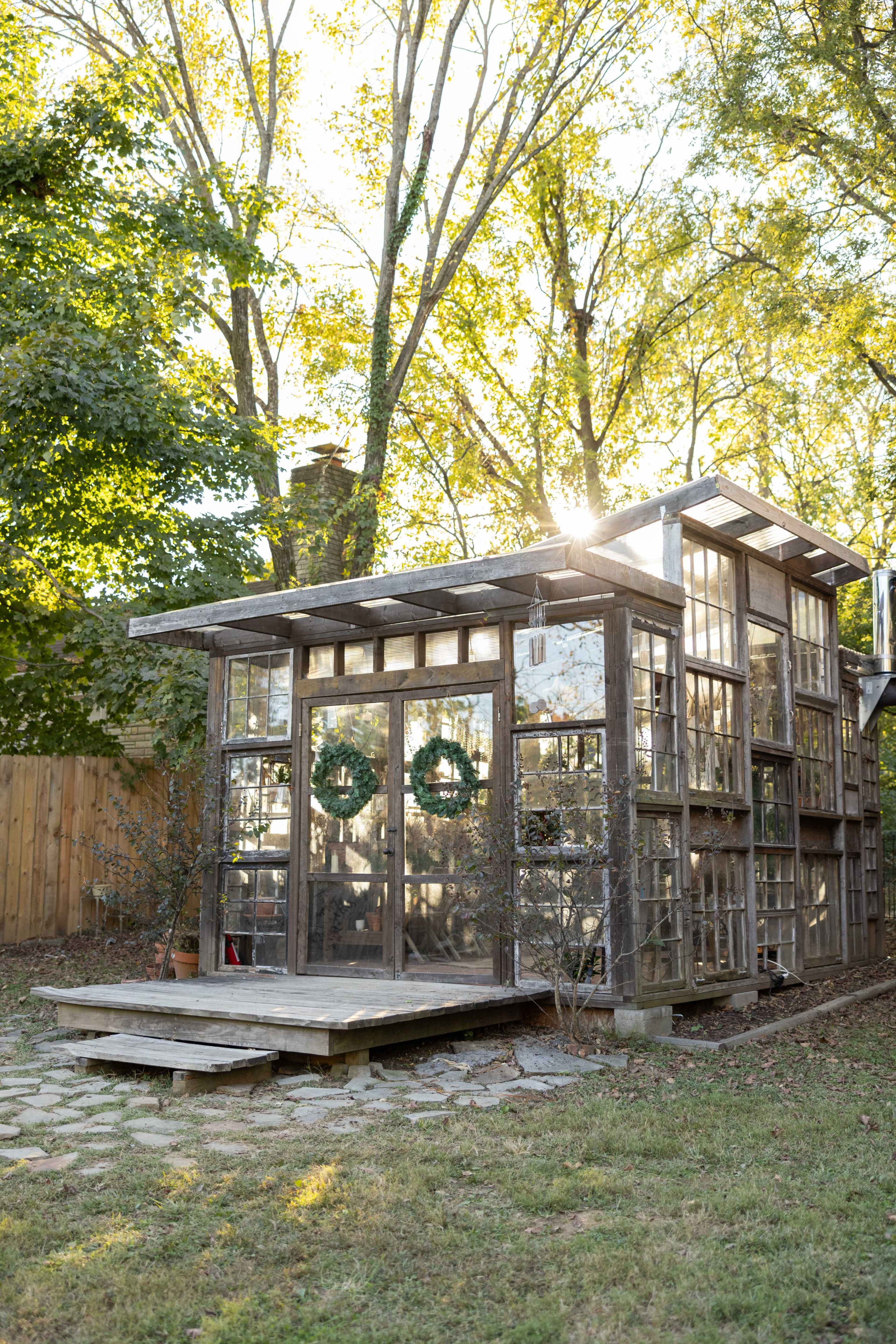 A rustic greenhouse made of reclaimed windows stands in a backyard, surrounded by trees and greenery.