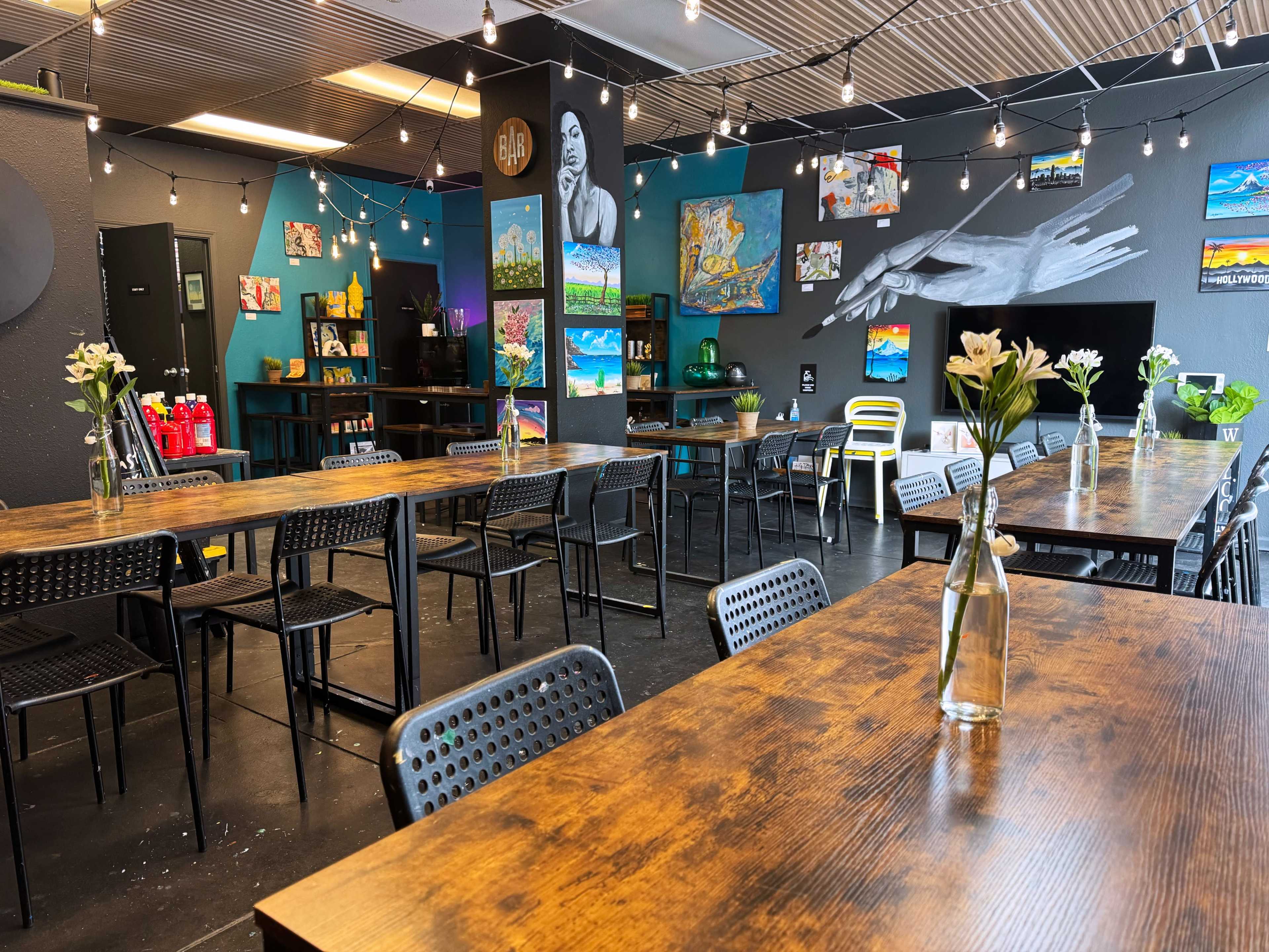 A spacious café with wooden tables and black chairs, decorated with colorful artwork on the walls and string lights overhead.