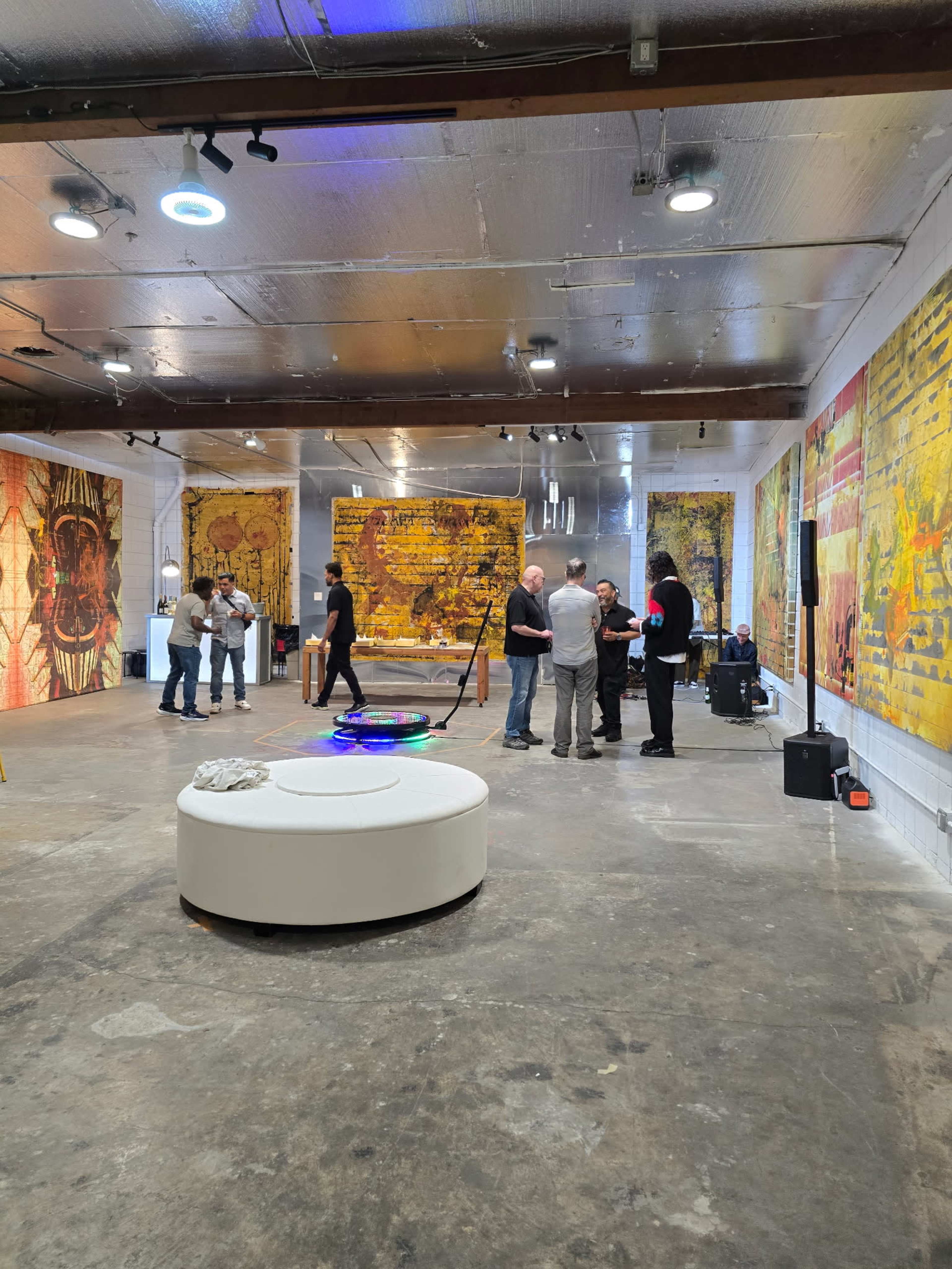 A group of people is gathered in an art gallery featuring large, vibrant murals on the walls and a circular white seating area in the center.