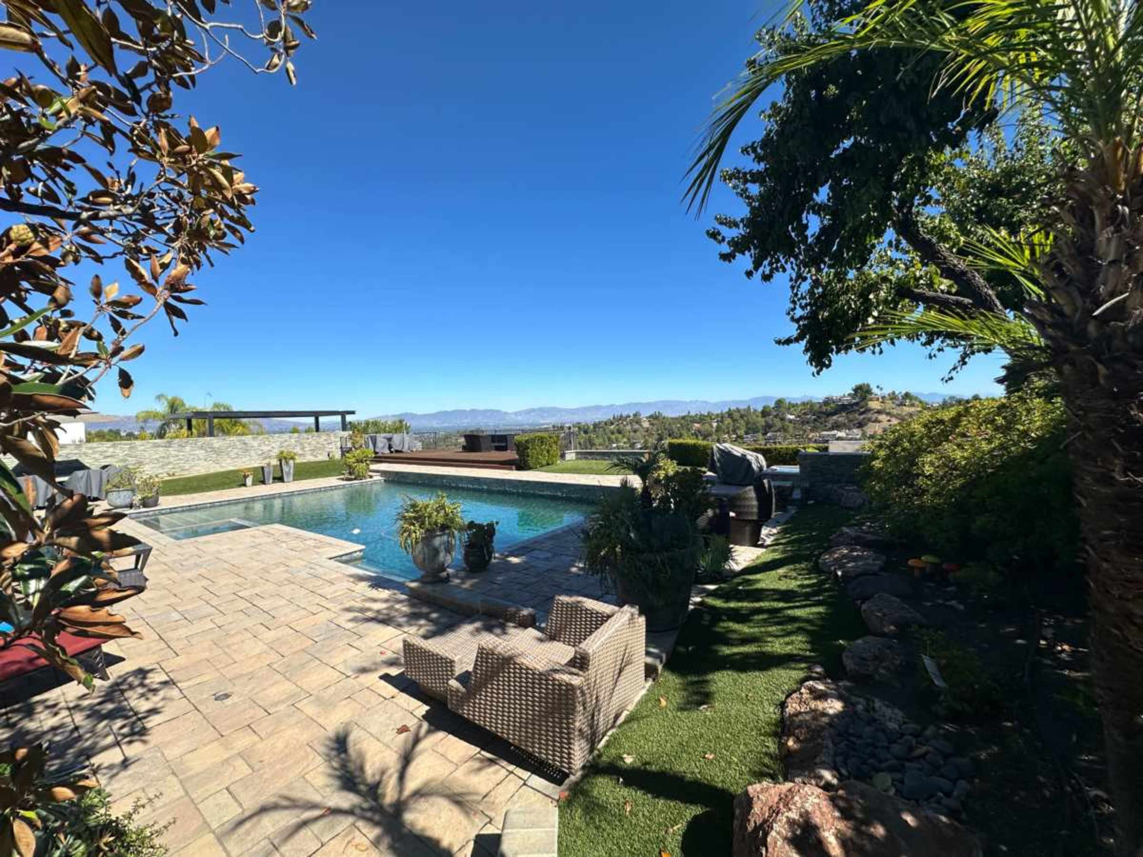 The image shows a landscaped backyard featuring a swimming pool surrounded by stone pavers, palm trees, and mountain views in the distance.