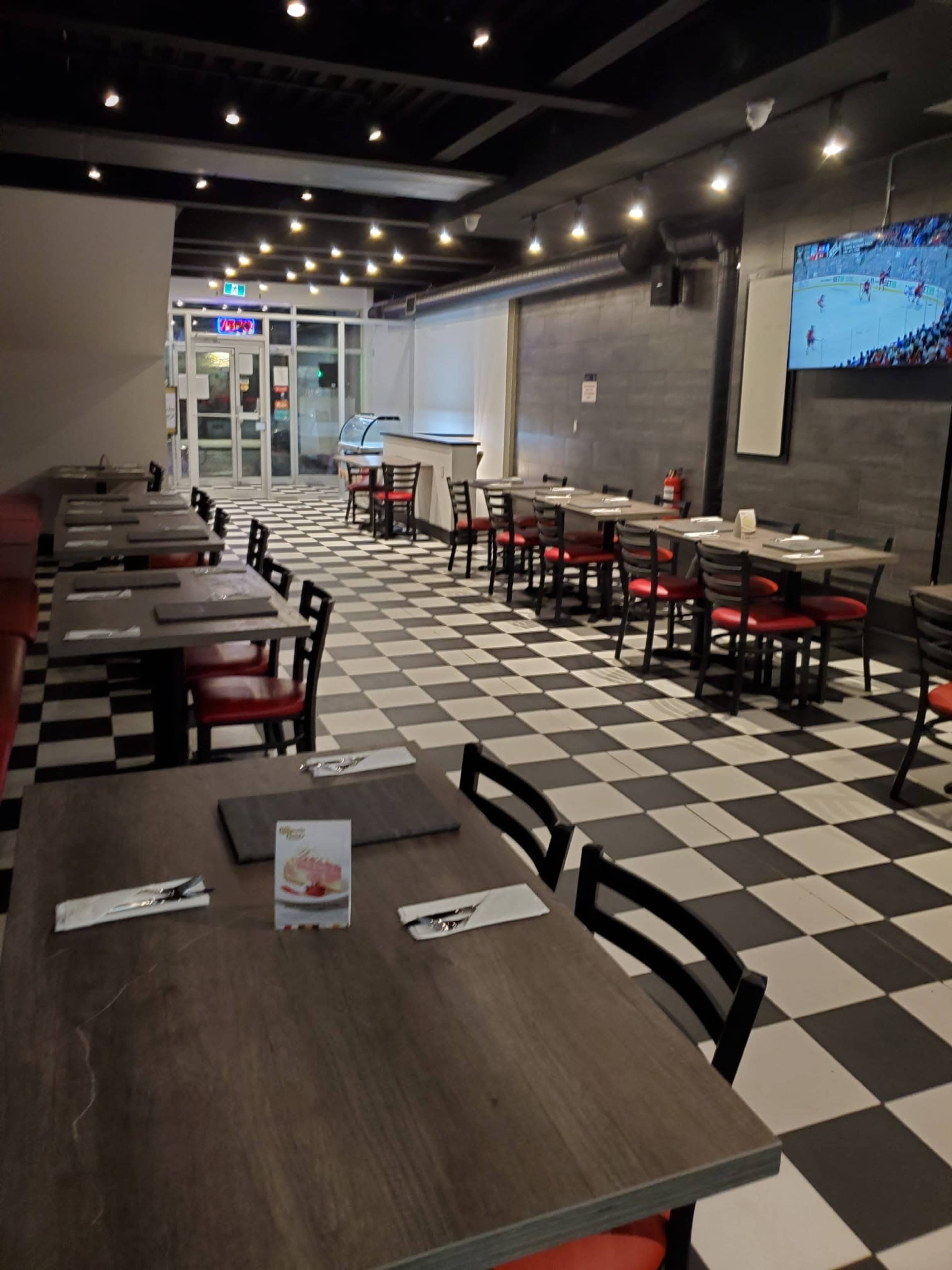 A mostly empty restaurant with black and white checkered flooring, red and black chairs, and a television displaying a sports game.