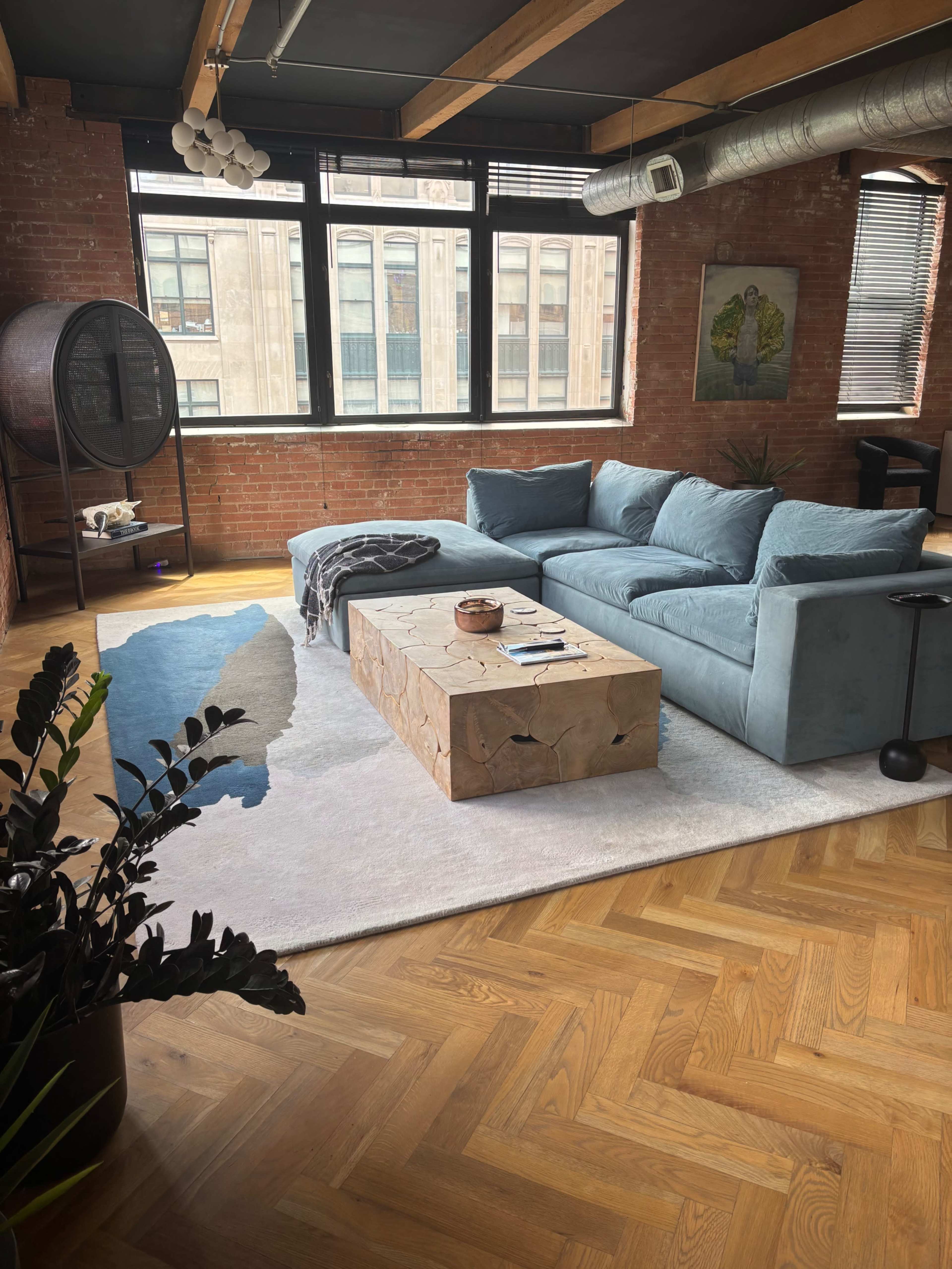 The image shows a modern living room featuring a blue sectional sofa, a wooden coffee table, a patterned area rug, and large windows with views of a brick building.