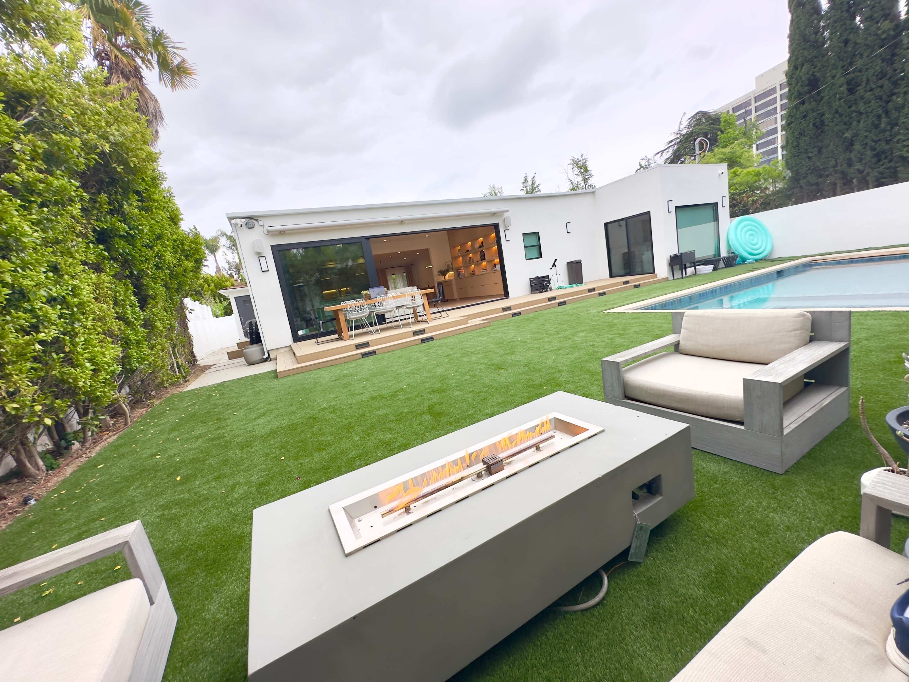 A modern backyard features a grassy area with a fire pit, lounge seating, and a pool, adjacent to a contemporary home with large windows.