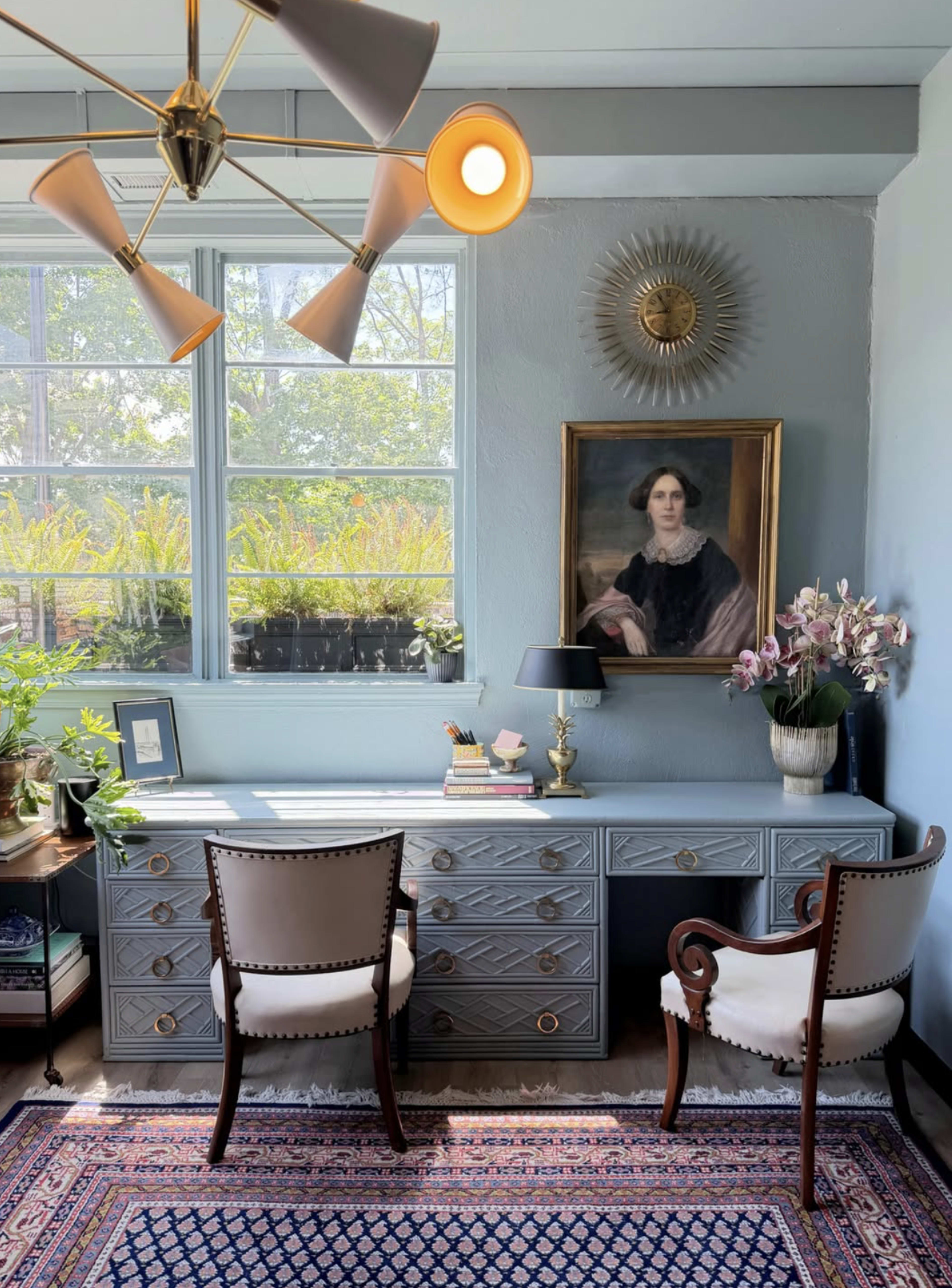 A well-decorated home office features a light blue desk with drawers, two decorative chairs, a wall-mounted portrait, a lamp, and a ceiling light fixture, all set against a backdrop of large windows with greenery outside.