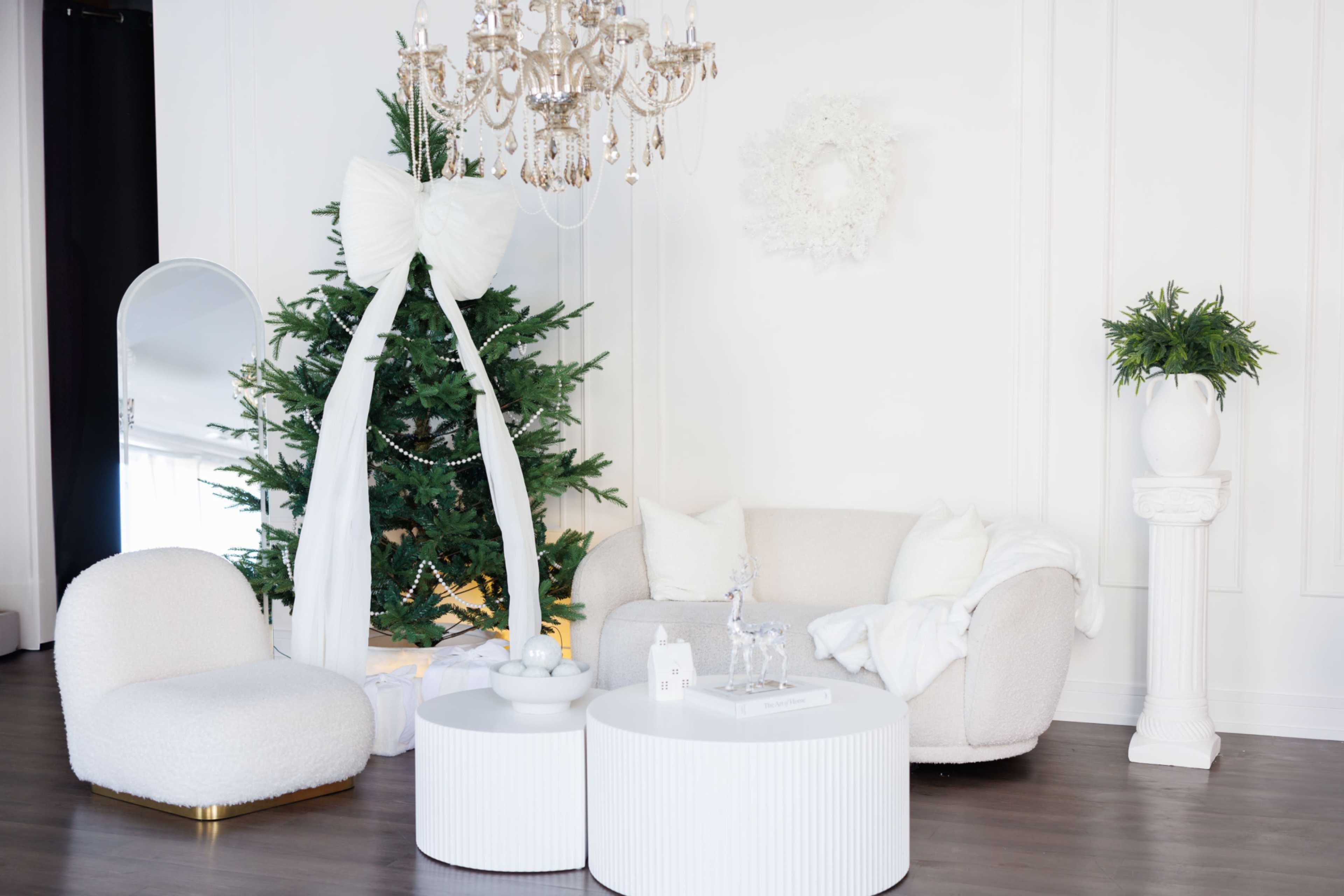 The image shows a decorated living room featuring a Christmas tree with a large white bow, a plush white sofa, and two round white coffee tables.