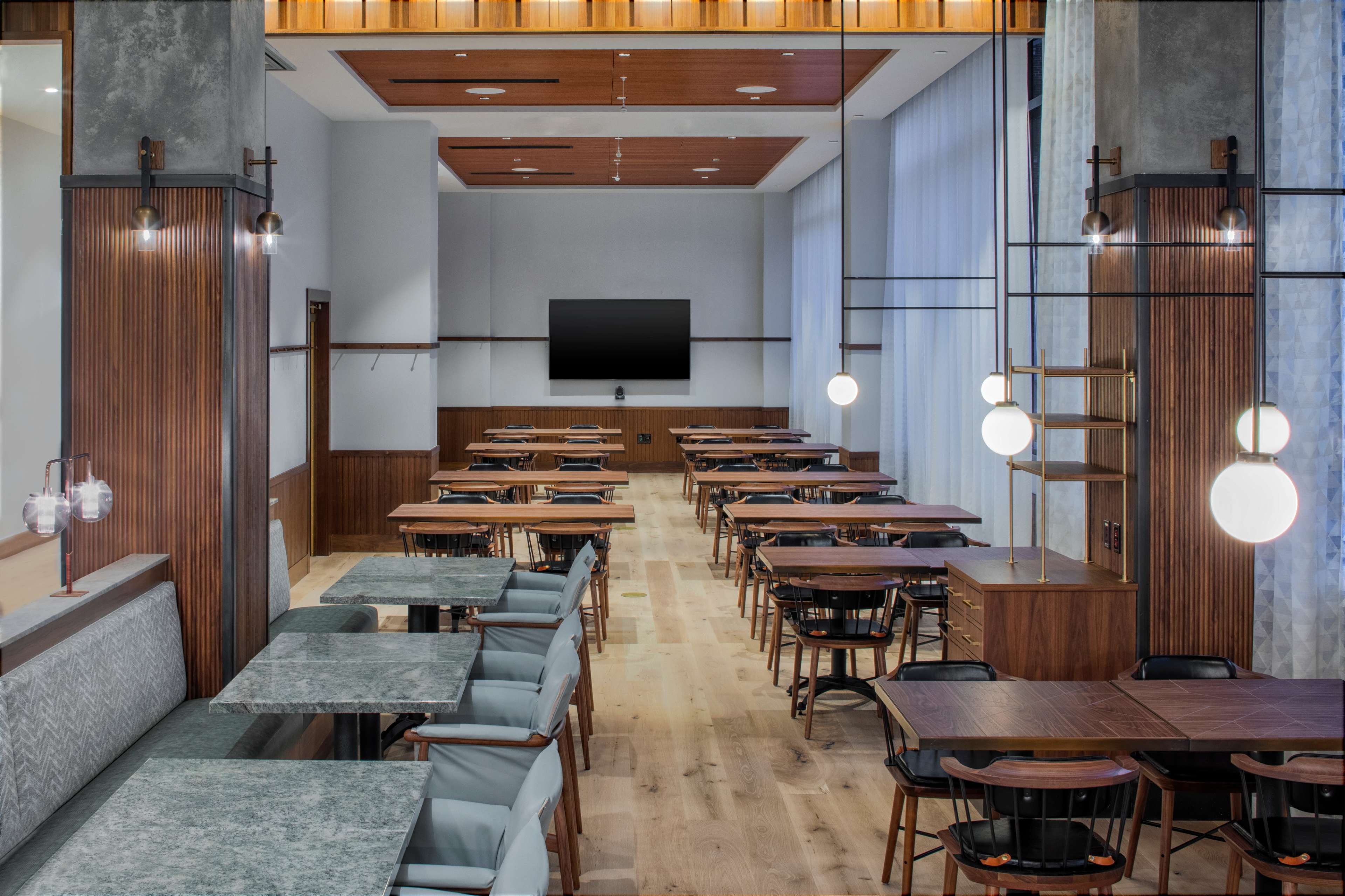 A modern restaurant interior featuring several rows of wooden tables and chairs, illuminated by pendant lights, with a large screen mounted on the wall.