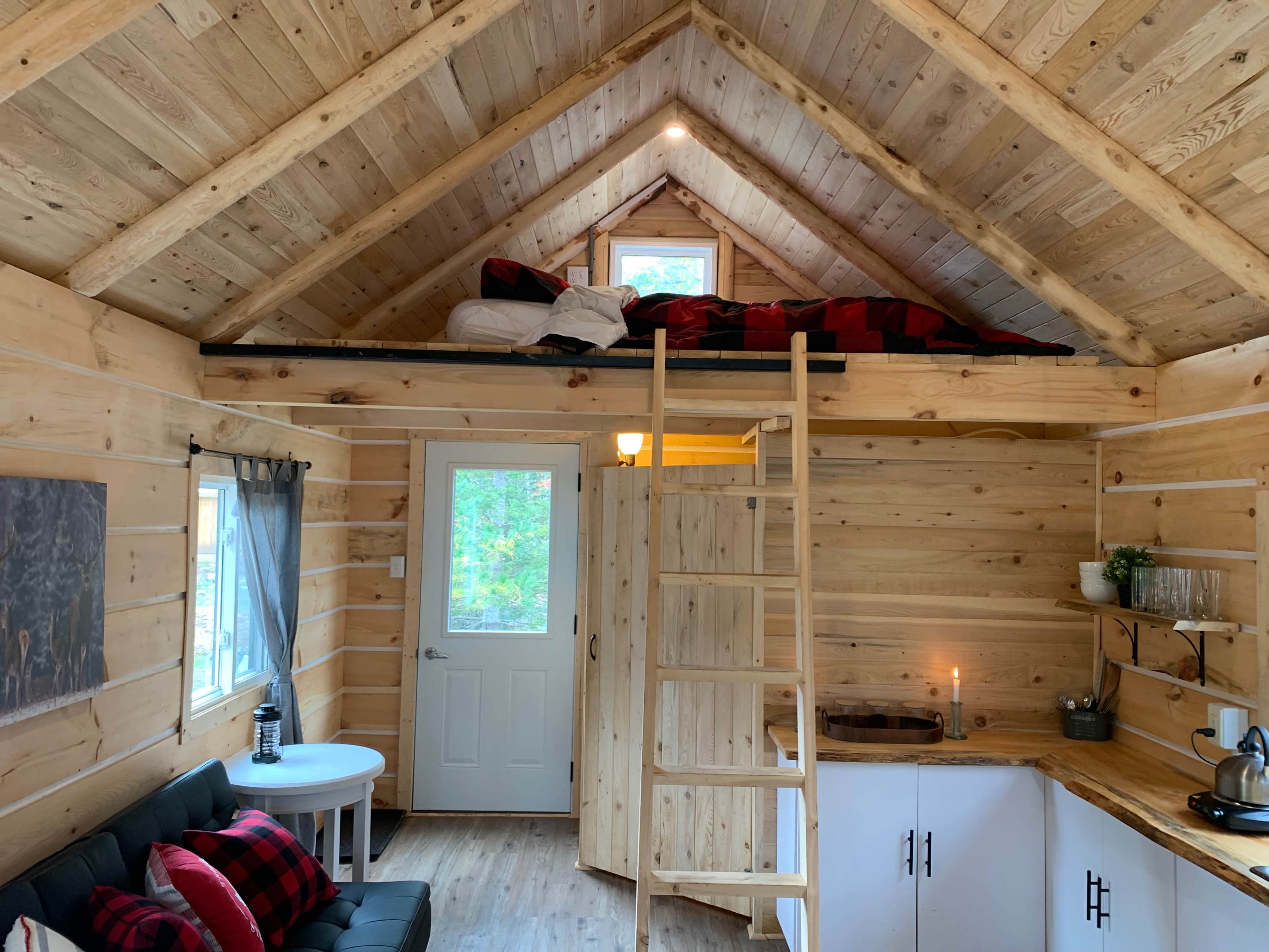 The image shows a cozy wooden interior of a small cabin with a lofted sleeping area, a ladder, and a kitchenette on one side.