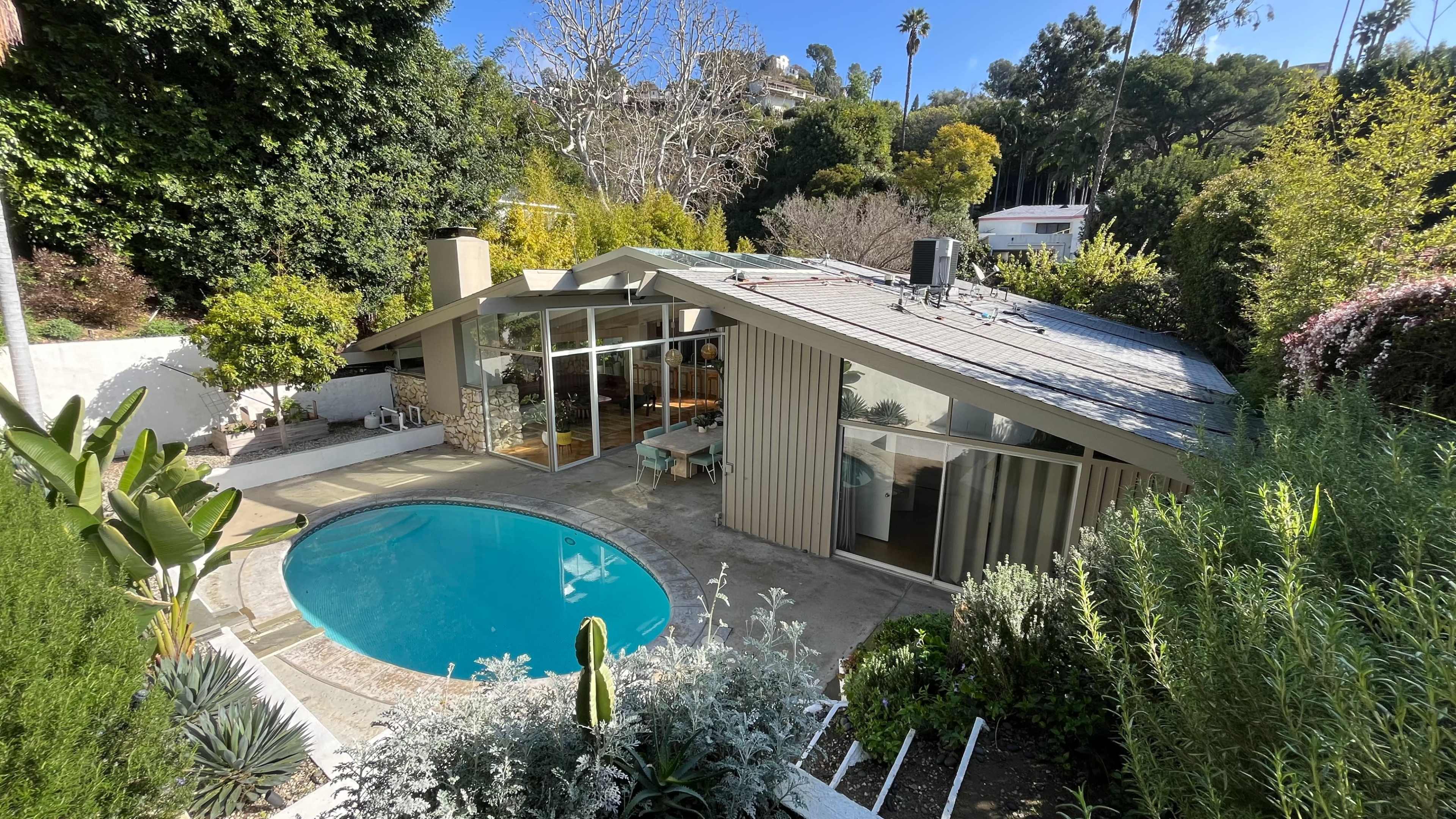 The image shows a modern house with large windows next to a circular swimming pool, surrounded by landscaped greenery.