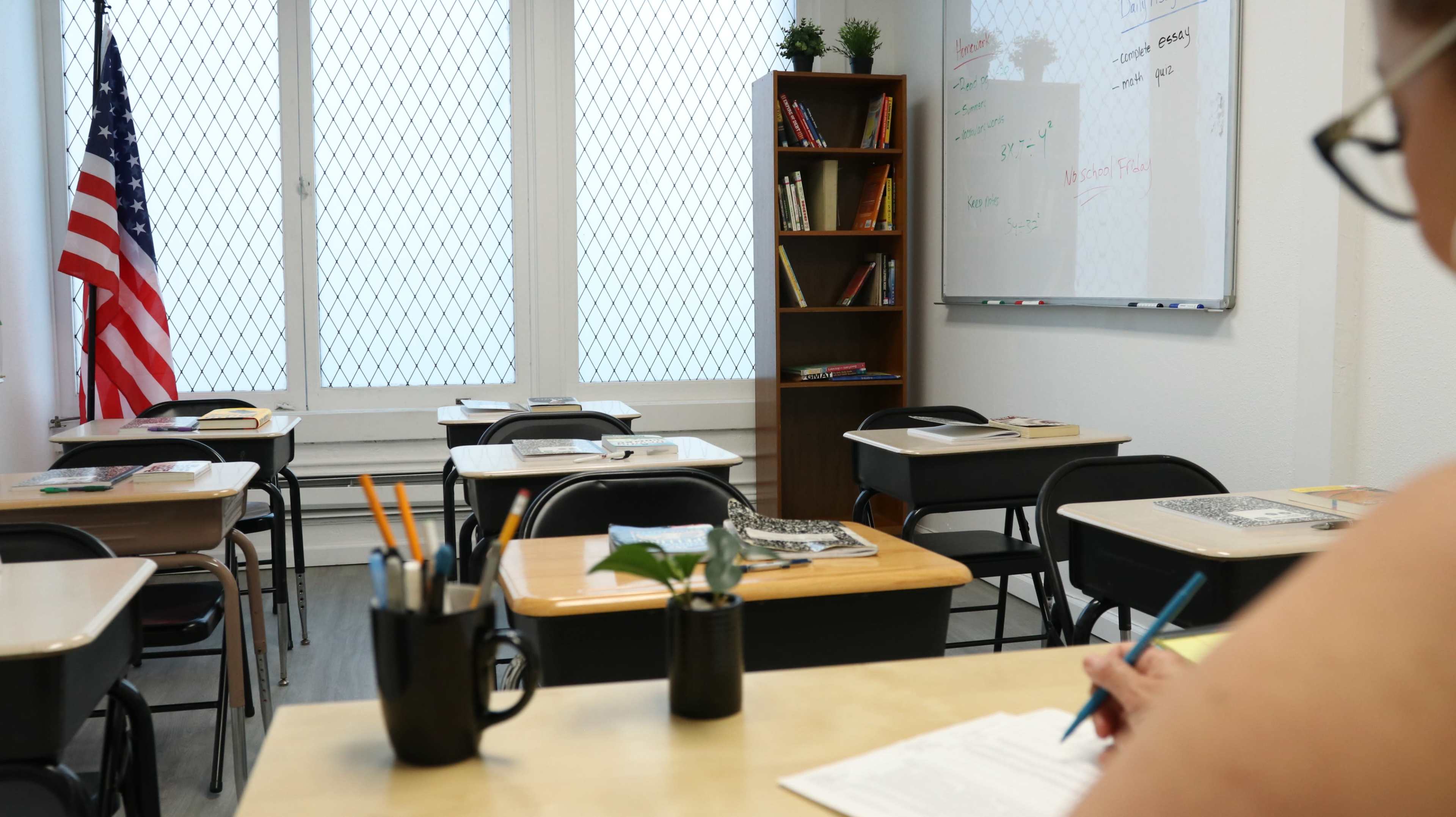 A classroom is shown with desks arranged in rows, a whiteboard at the front, and a bookshelf filled with books.