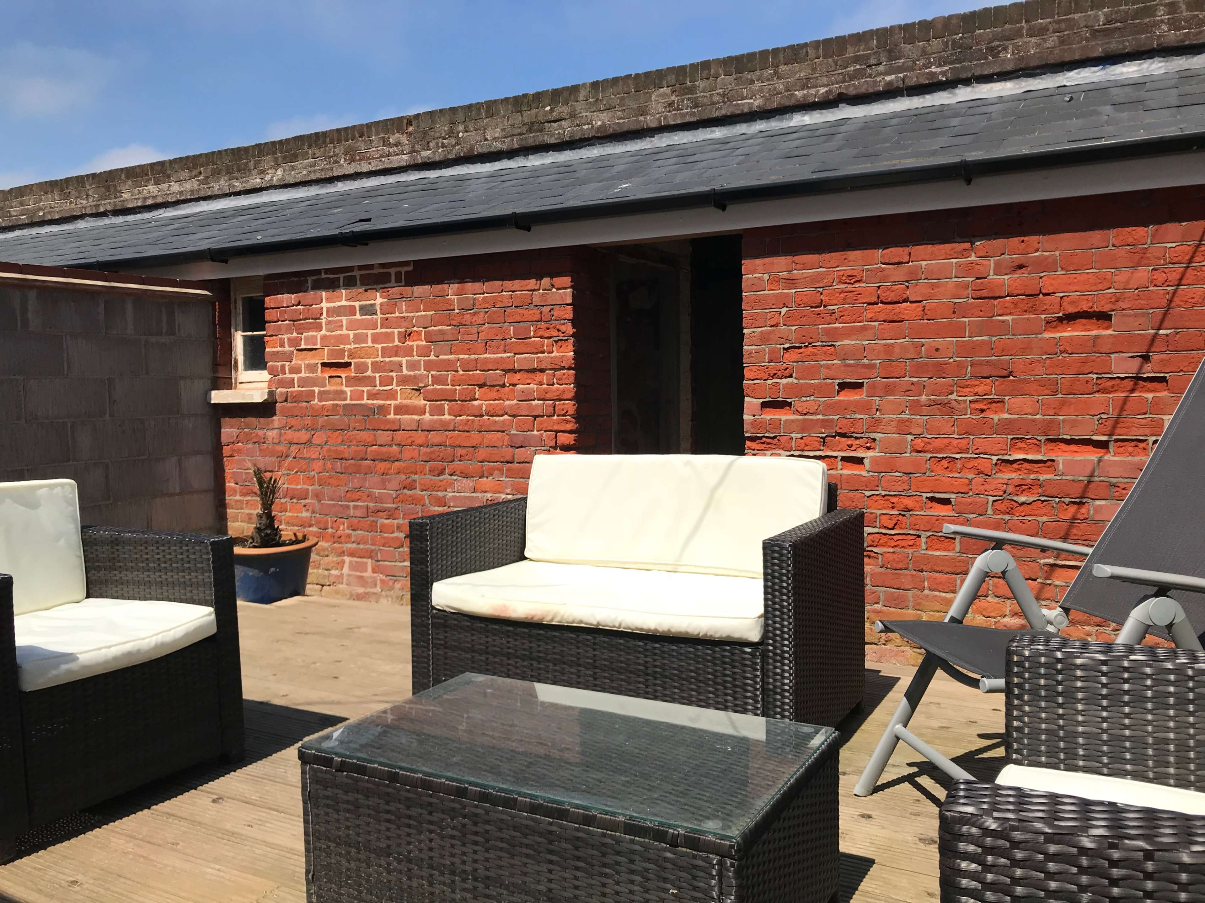 A patio area with outdoor furniture, including two black wicker chairs and a glass-top table, set against a brick wall and a blue sky.