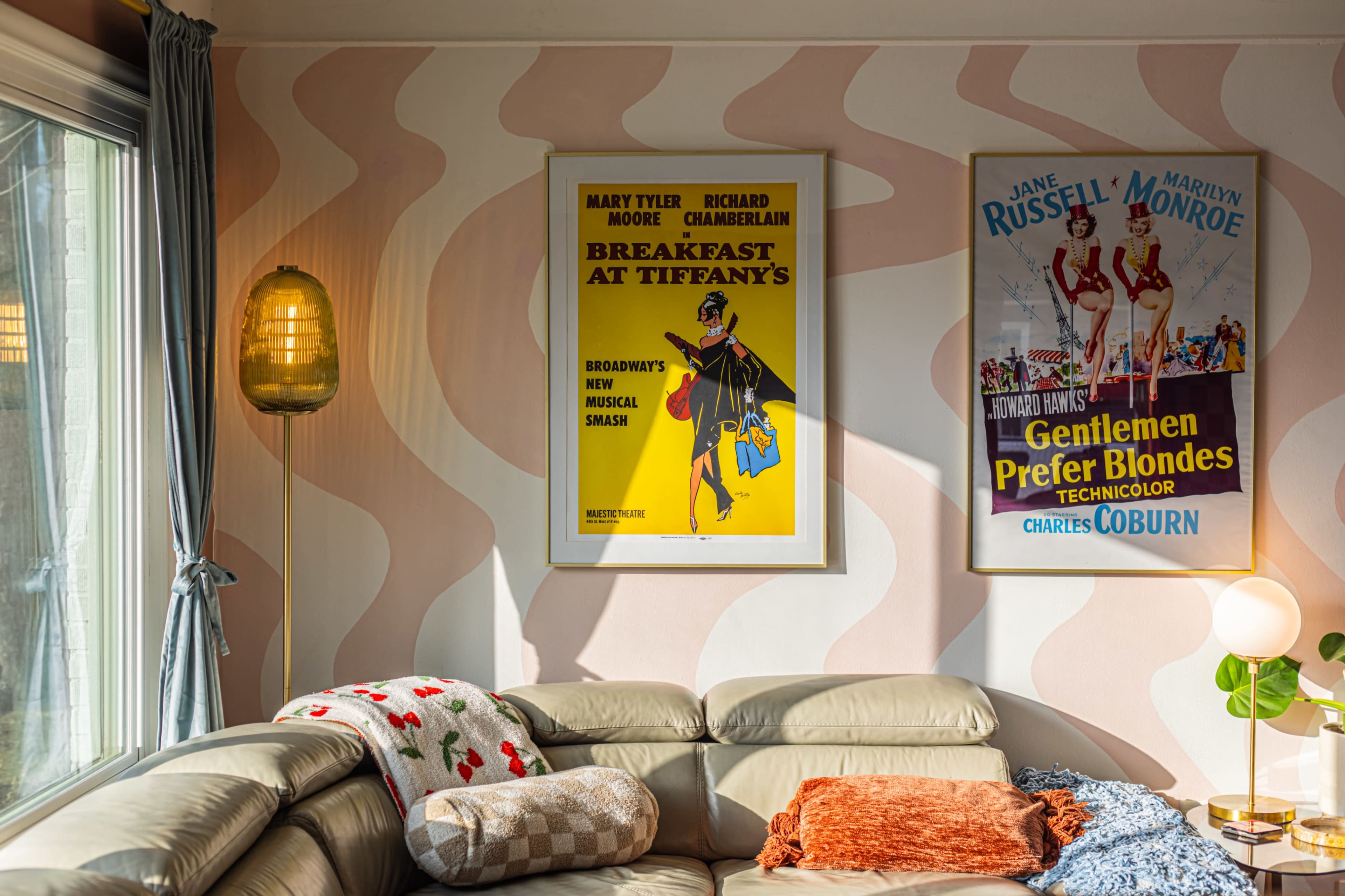 The image features a cozy living room with soft pink walls, two vintage movie posters framed on the wall, and a light-colored sofa adorned with decorative cushions.