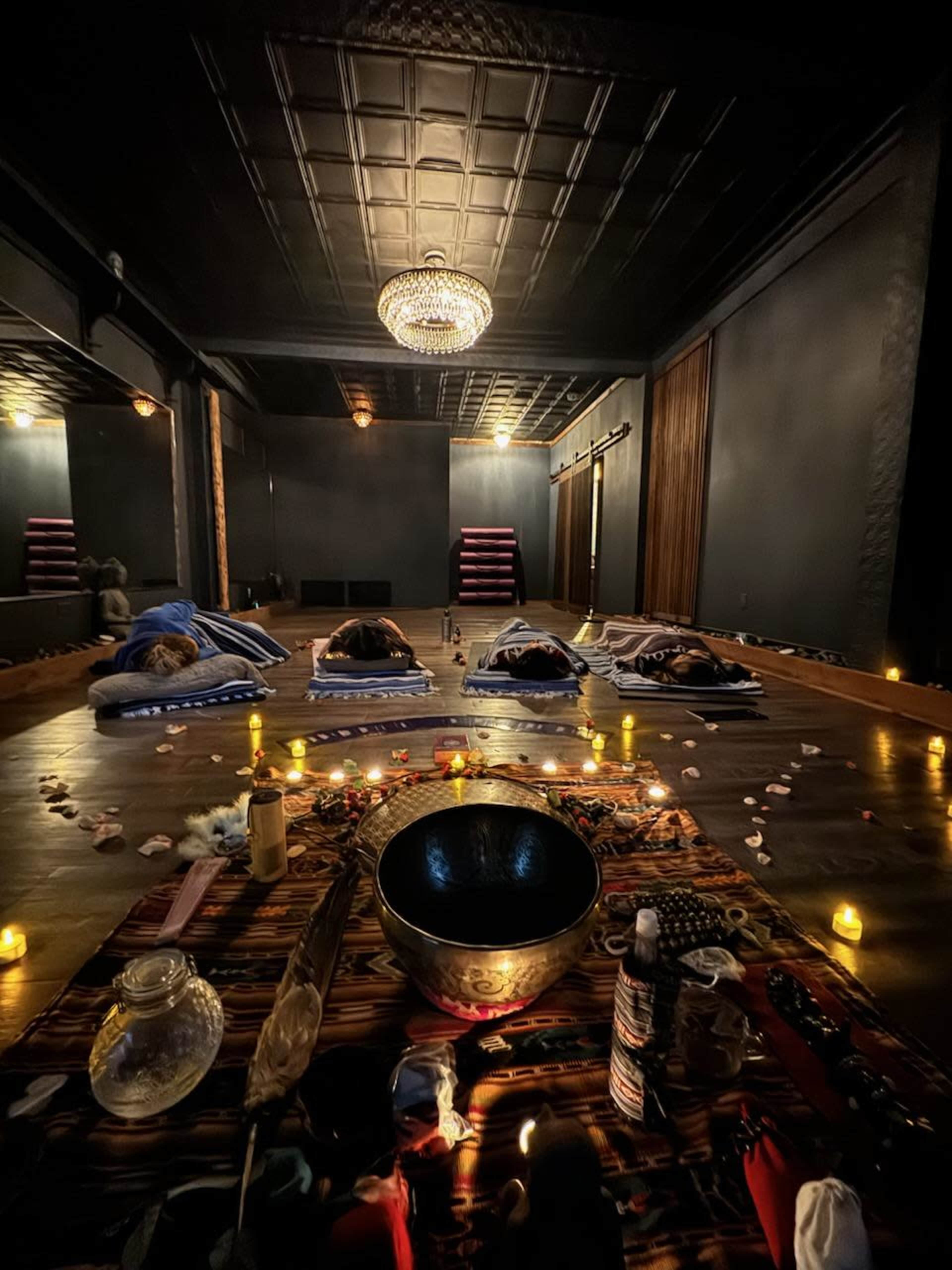 A dimly lit meditation room with several mattresses arranged on the wooden floor, surrounded by candles and a bowl at the center.