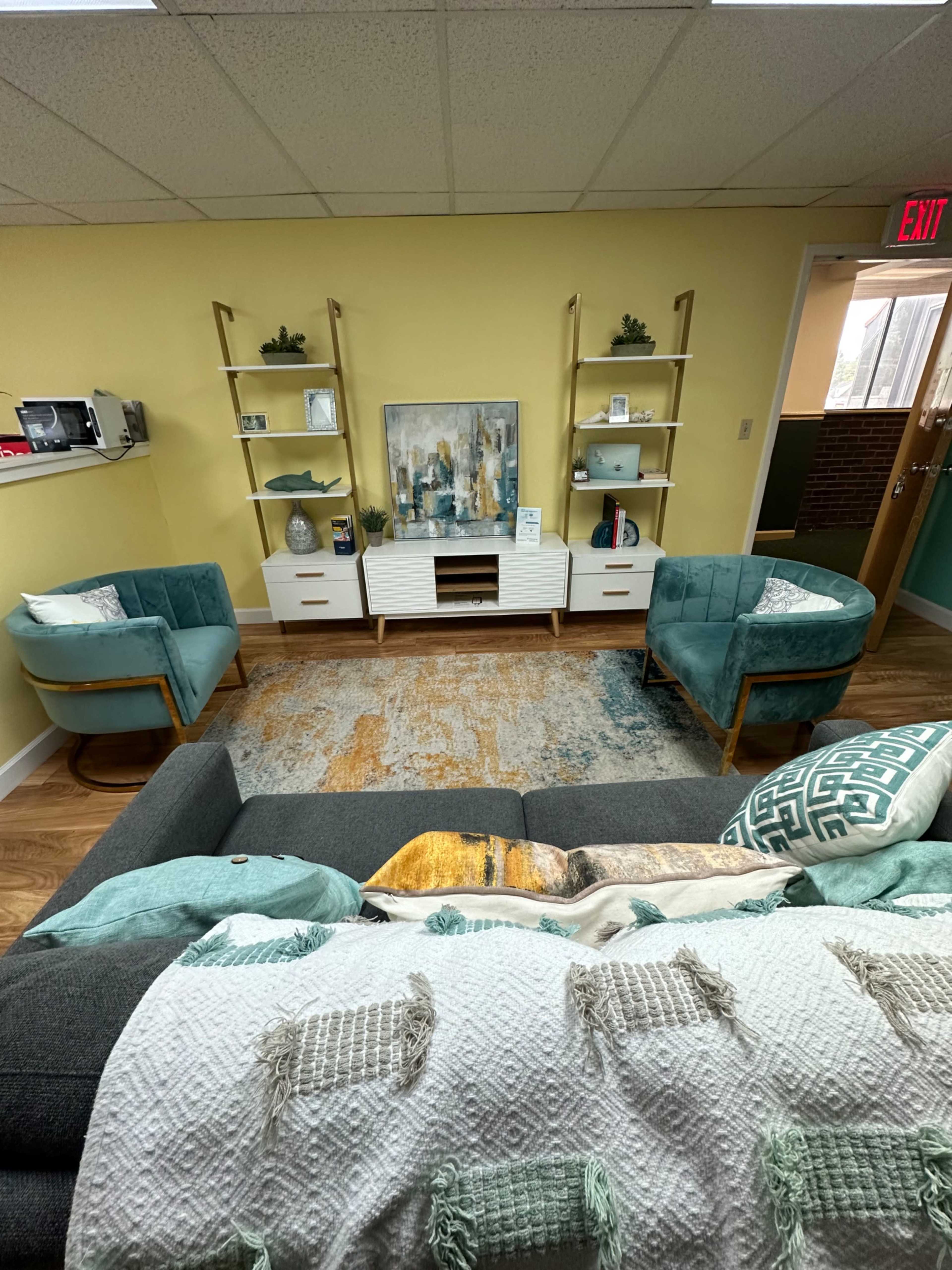 A modern living area featuring a gray sofa, two teal armchairs, a rug, a media console with shelves, and a decorative wall painting.