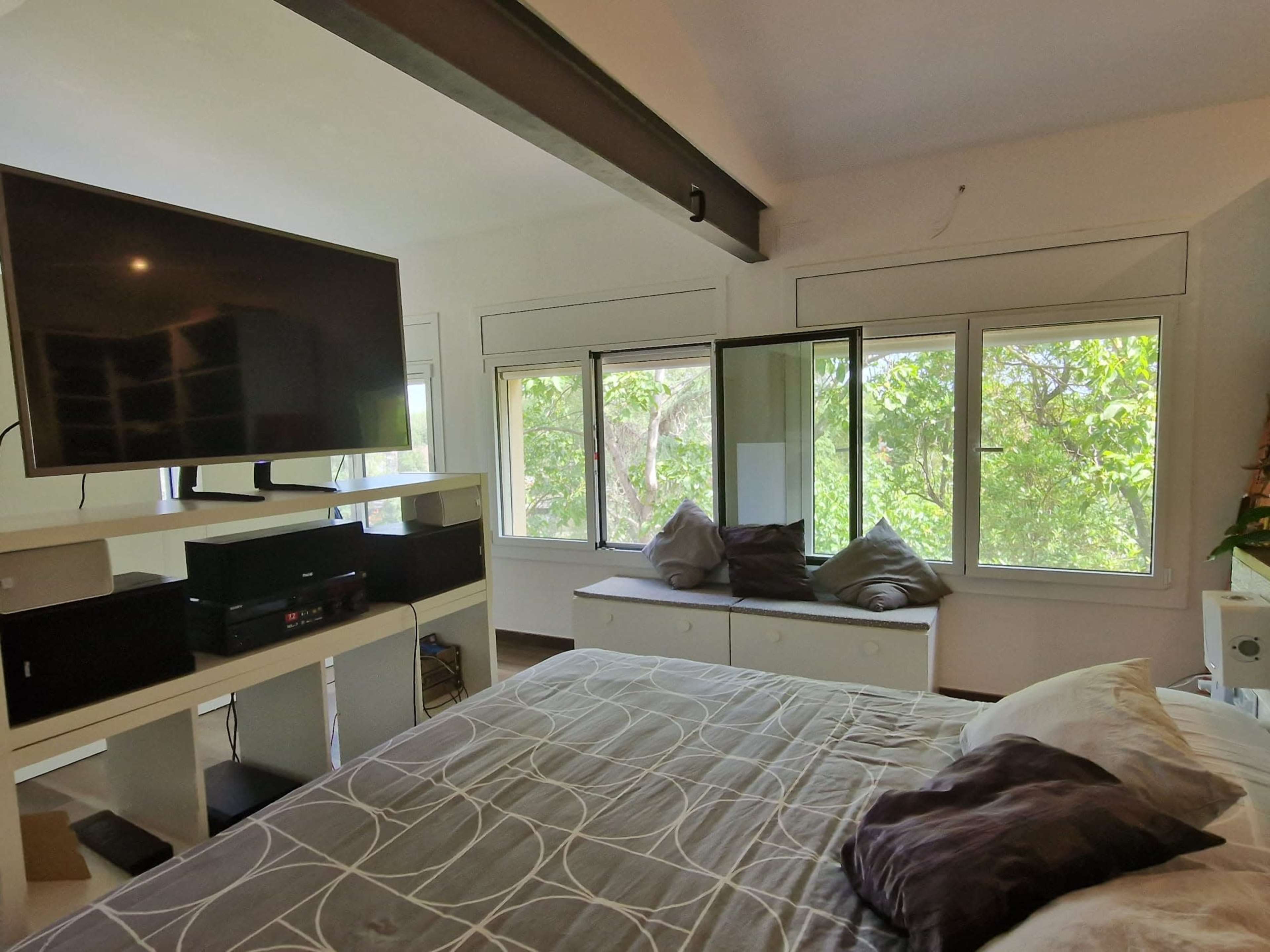 A modern bedroom features a large television mounted on the wall, a bed with gray bedding, and multiple windows offering a view of greenery outside.