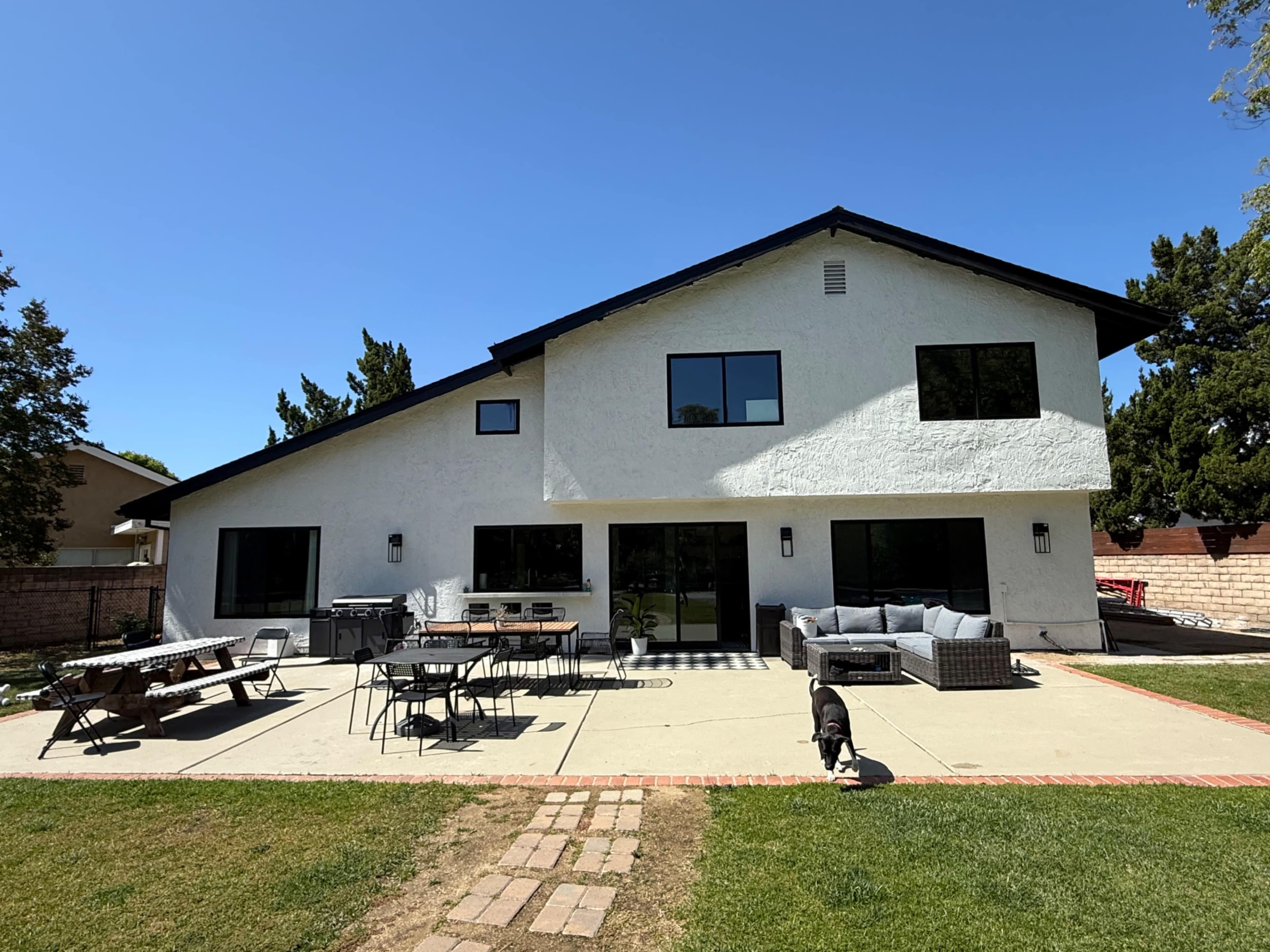 A white two-story house with large windows features a patio area with outdoor furniture and a grassy lawn.