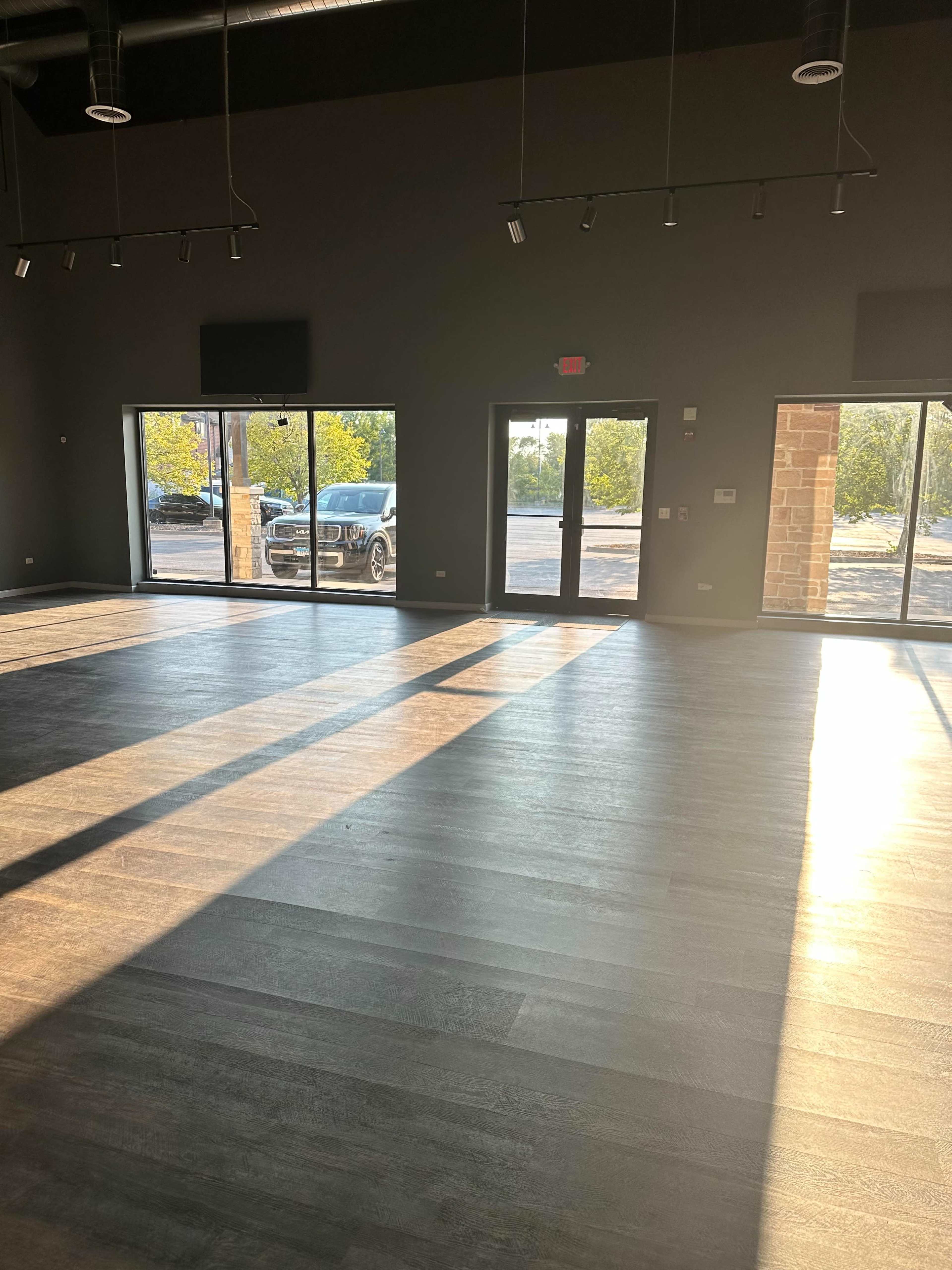 The image shows a spacious, empty room with large windows allowing sunlight to stream in, casting long shadows across the wooden floor.