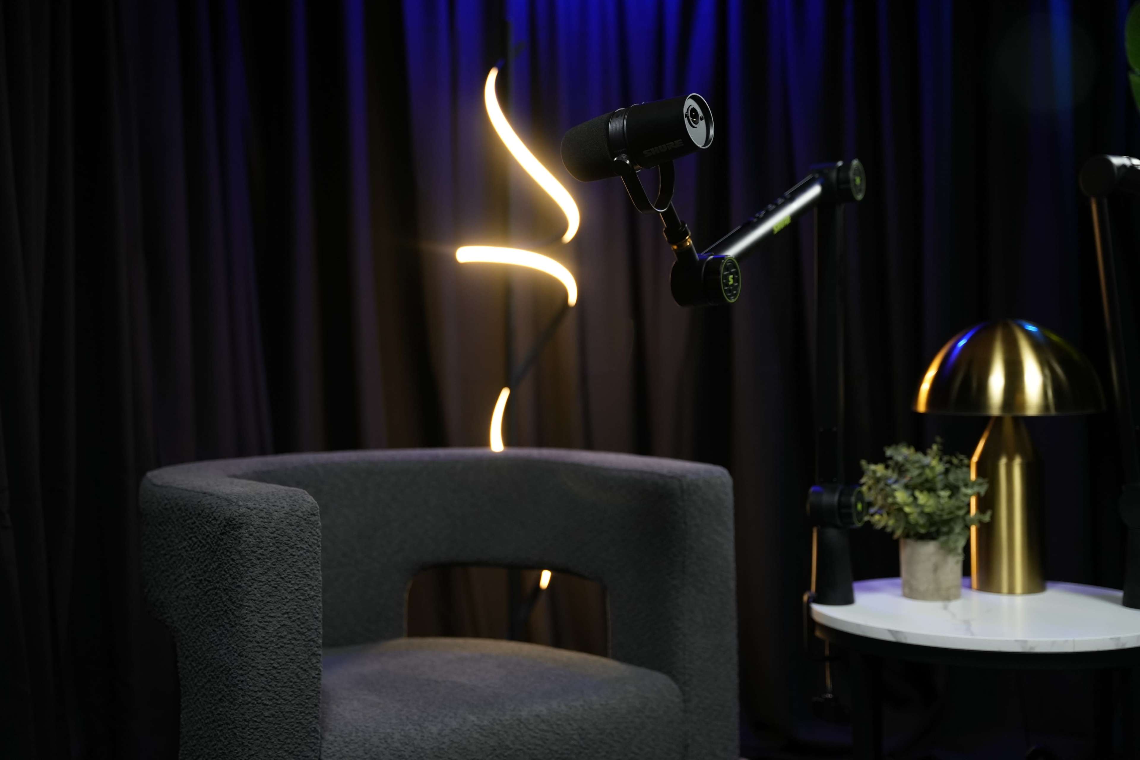 The image features a gray armchair beside a small white table with a decorative plant and a lamp, set against a backdrop of dark curtains and a glowing light fixture.