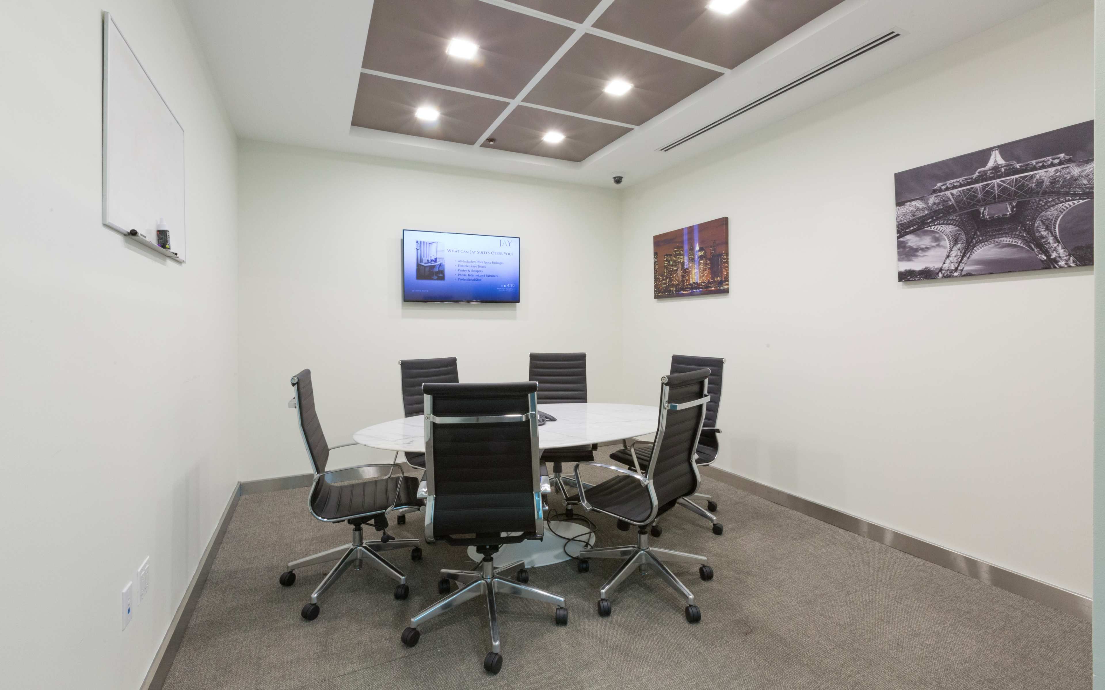 A conference room features a round table surrounded by six black office chairs, a wall-mounted screen, and framed cityscape photographs.