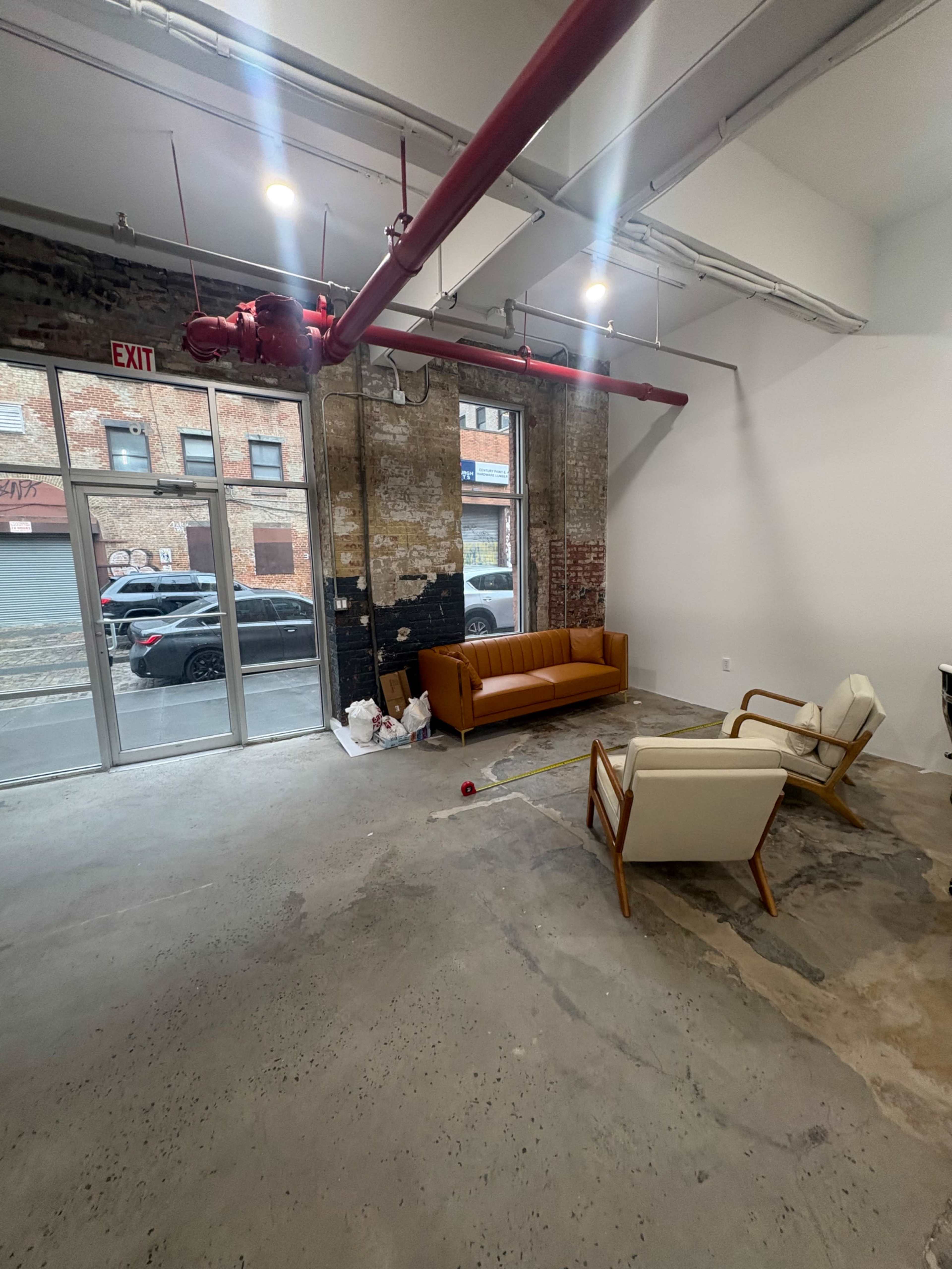 The image shows a spacious, minimally furnished room with a brown leather sofa and two white armchairs, featuring exposed brick walls and industrial-style lighting.