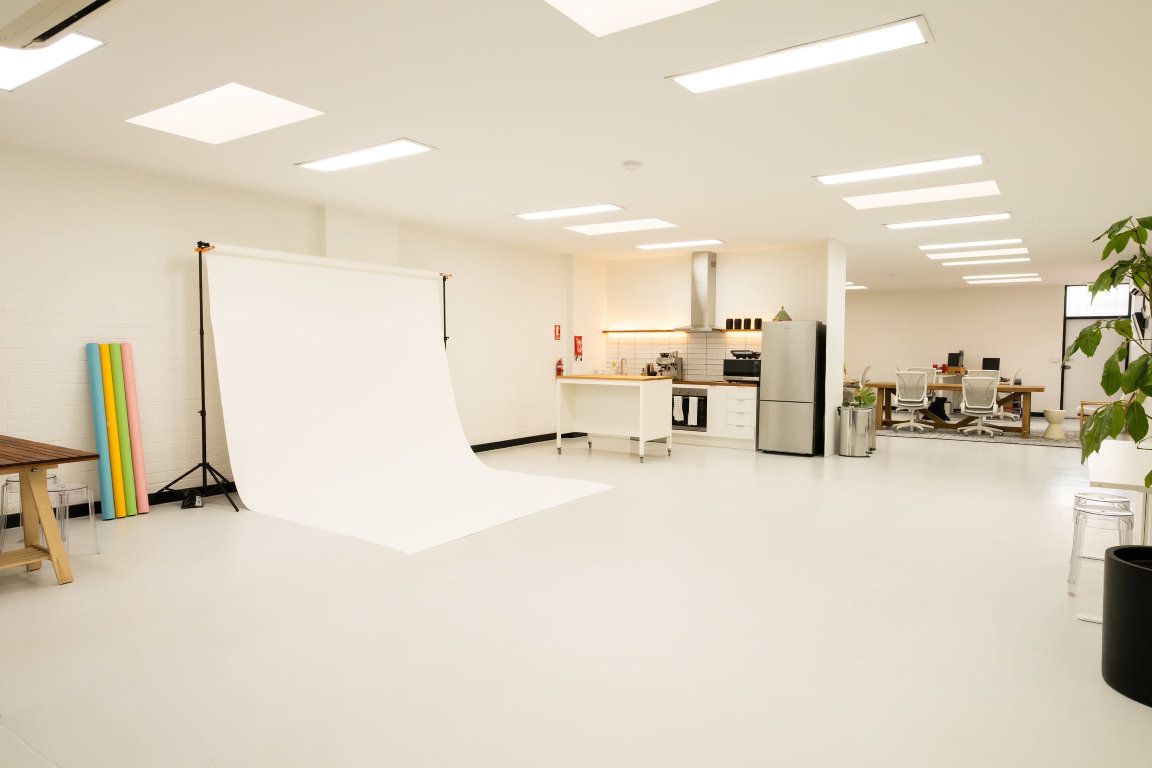 The image shows a bright, minimalist studio space with a white backdrop, a kitchenette, and modern office furniture.