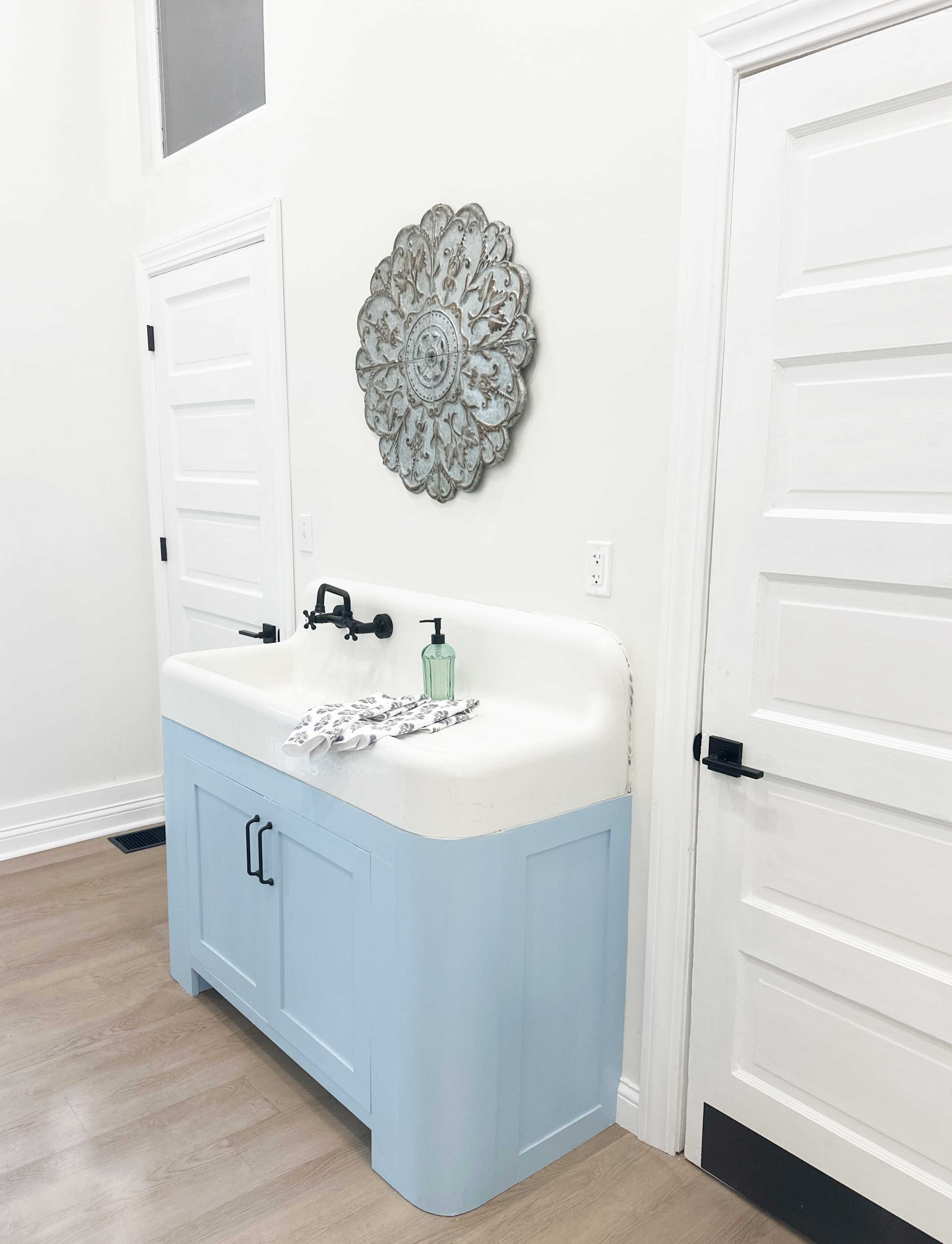 The image shows a light blue sink cabinet with a white countertop, a decorative wall piece above, and two white doors on either side in a bright room.