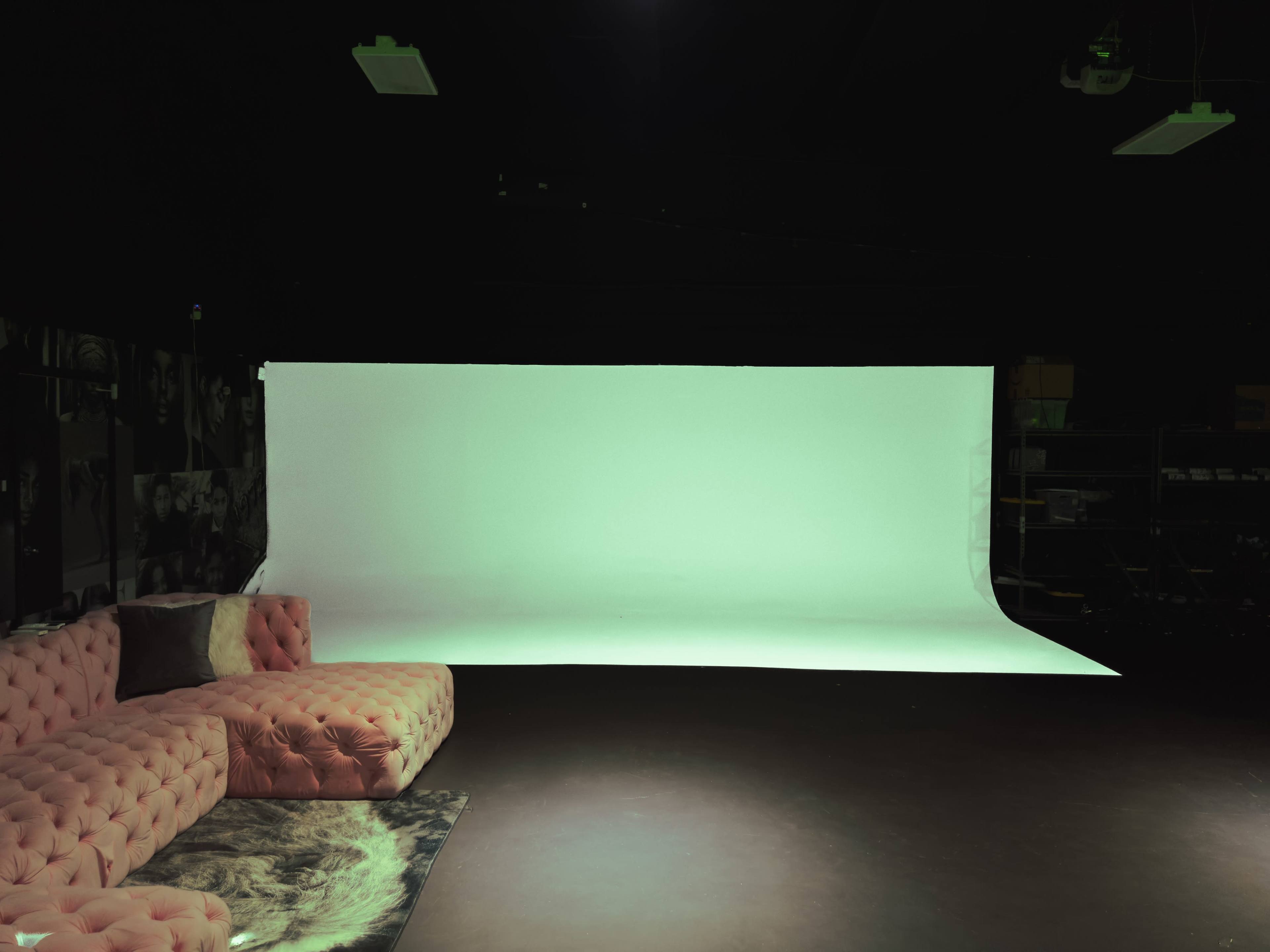 A studio space featuring a pink velvet sectional sofa, a patterned rug, and a large white backdrop.