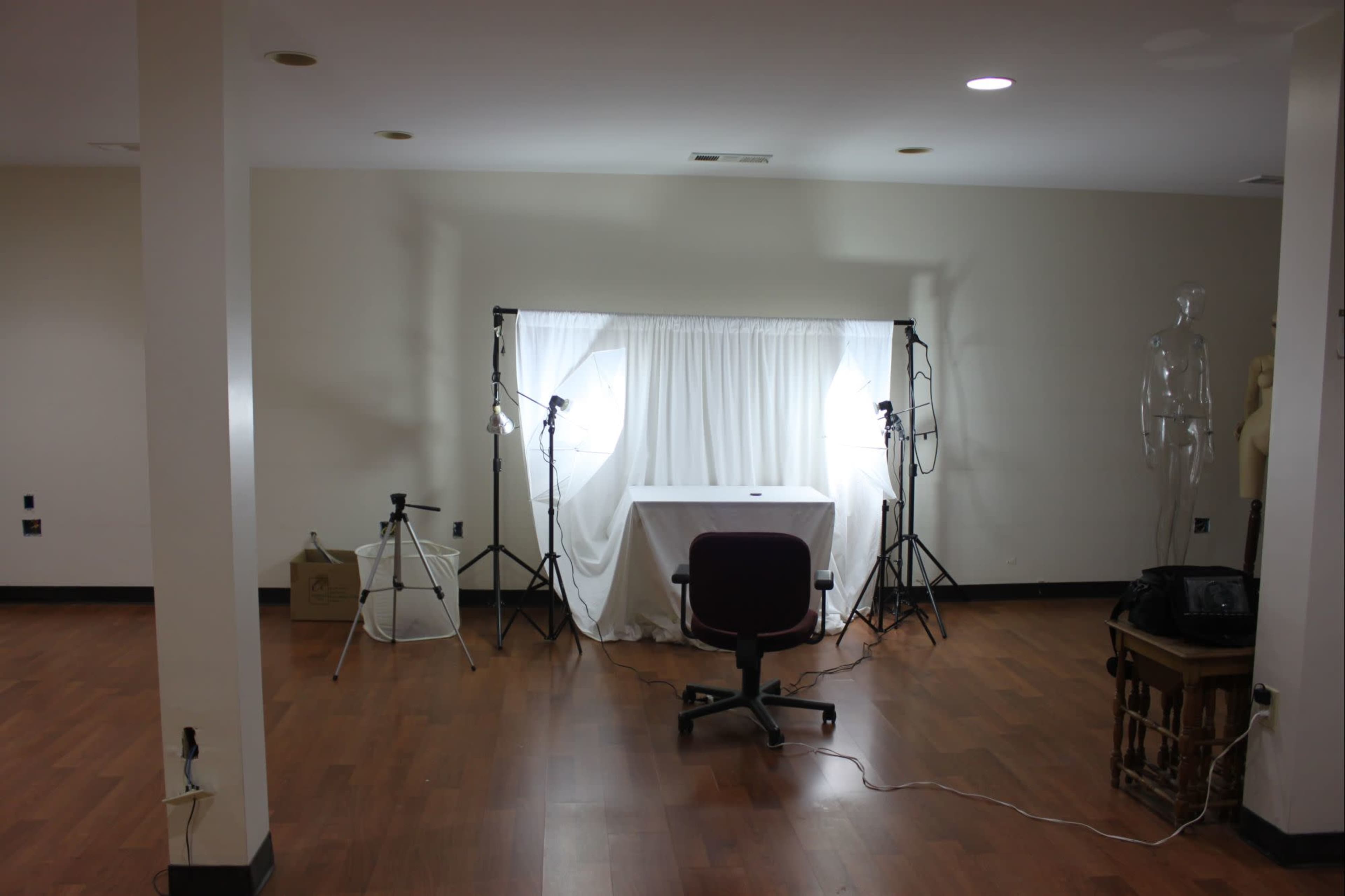 The image shows a spacious room with a table covered by a white cloth, flanked by two light stands and a chair, with a mannequin in the background.