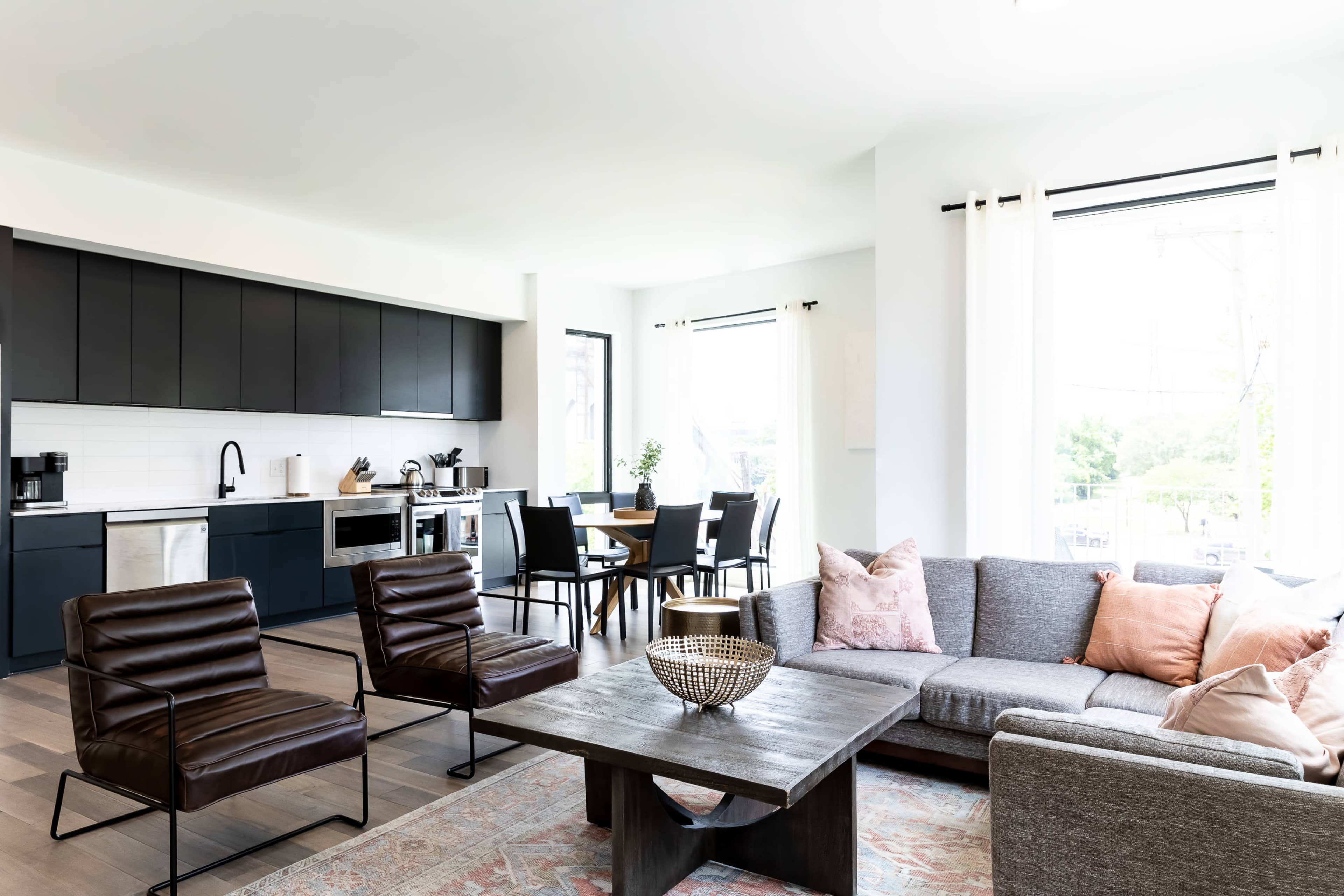 A modern living space features a gray sofa and brown chairs, a wooden coffee table, and a kitchen visible in the background with dark cabinetry.