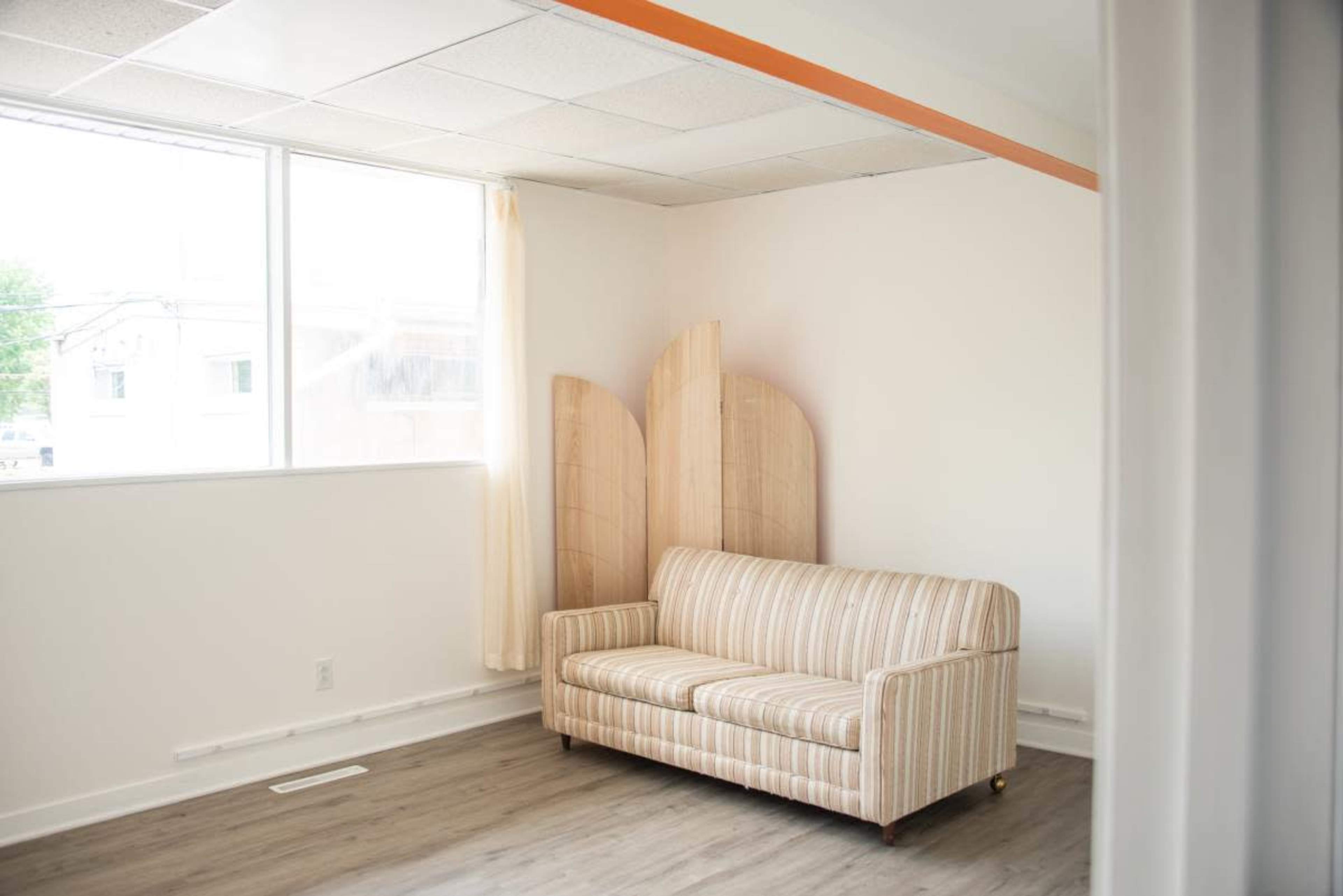 The image shows a light and airy room featuring a striped sofa, a large window with a curtain, and wooden panels in the background.