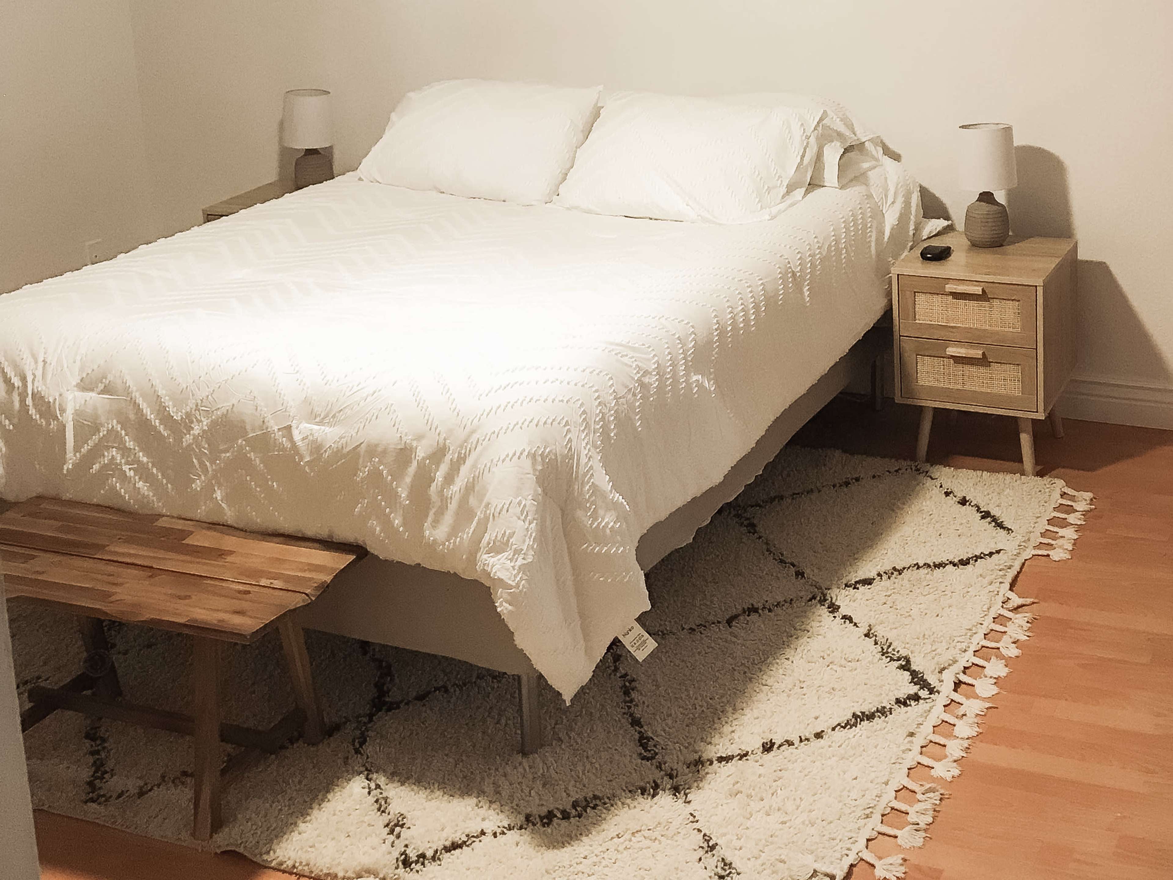 A bed with a white blanket is positioned against the wall, flanked by two nightstands with lamps, and there is a wooden bench at the foot of the bed on a patterned rug.
