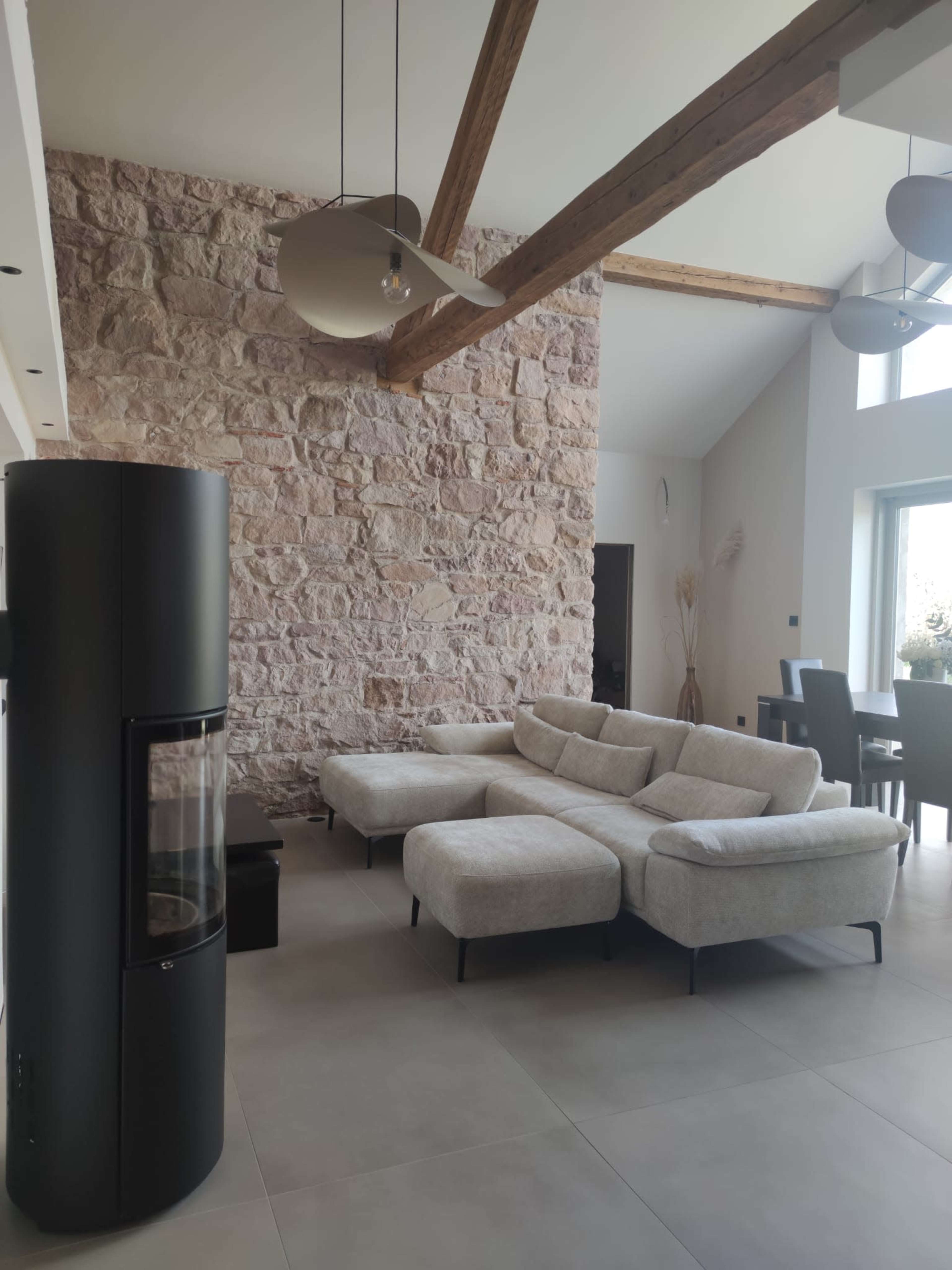 The image shows a modern living room featuring a light-colored sectional sofa, a sleek black fireplace, exposed wooden beams, and a stone accent wall.