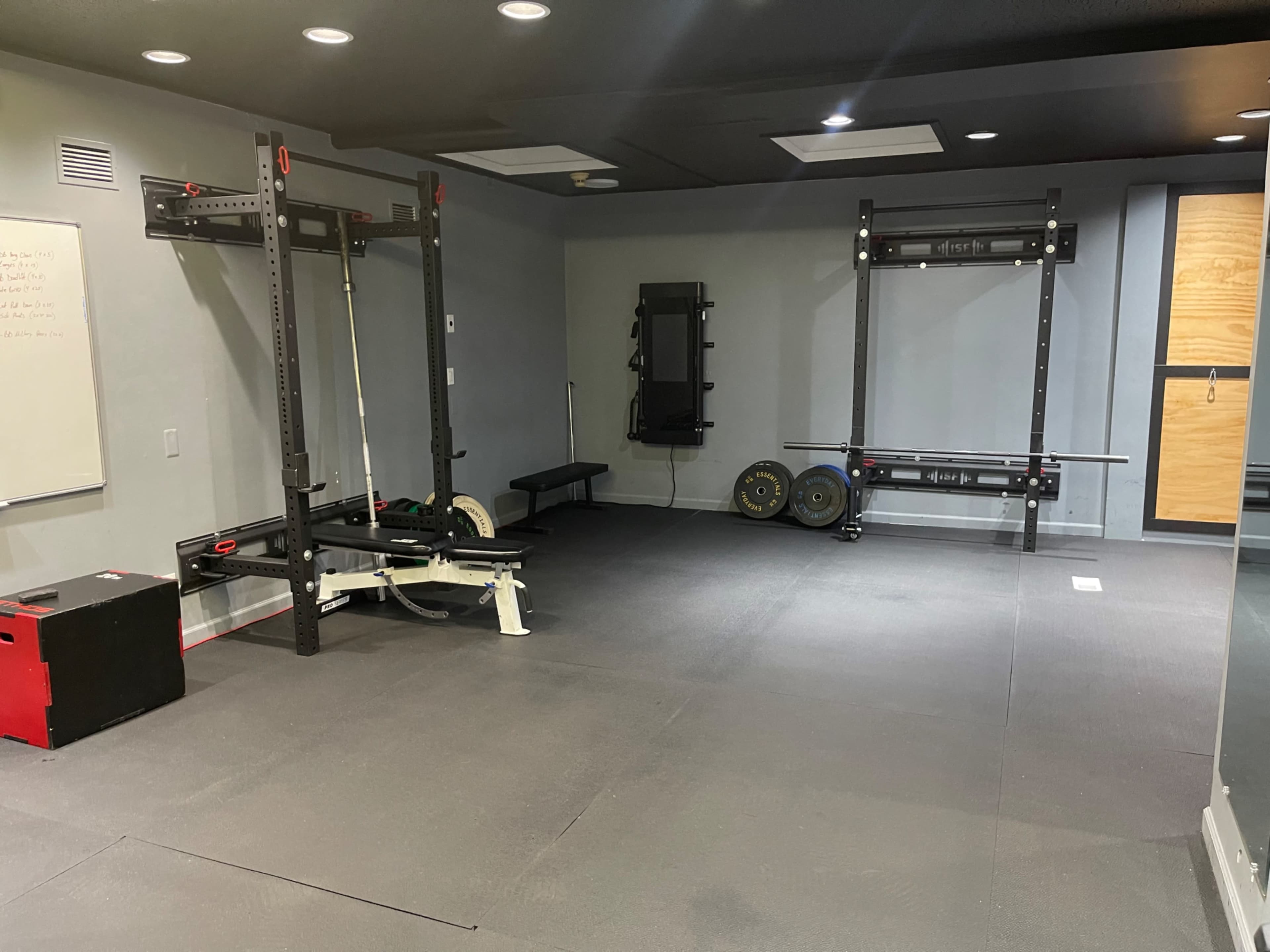 A minimalist home gym equipped with weightlifting racks, a bench, and exercise equipment on a rubber flooring surface.
