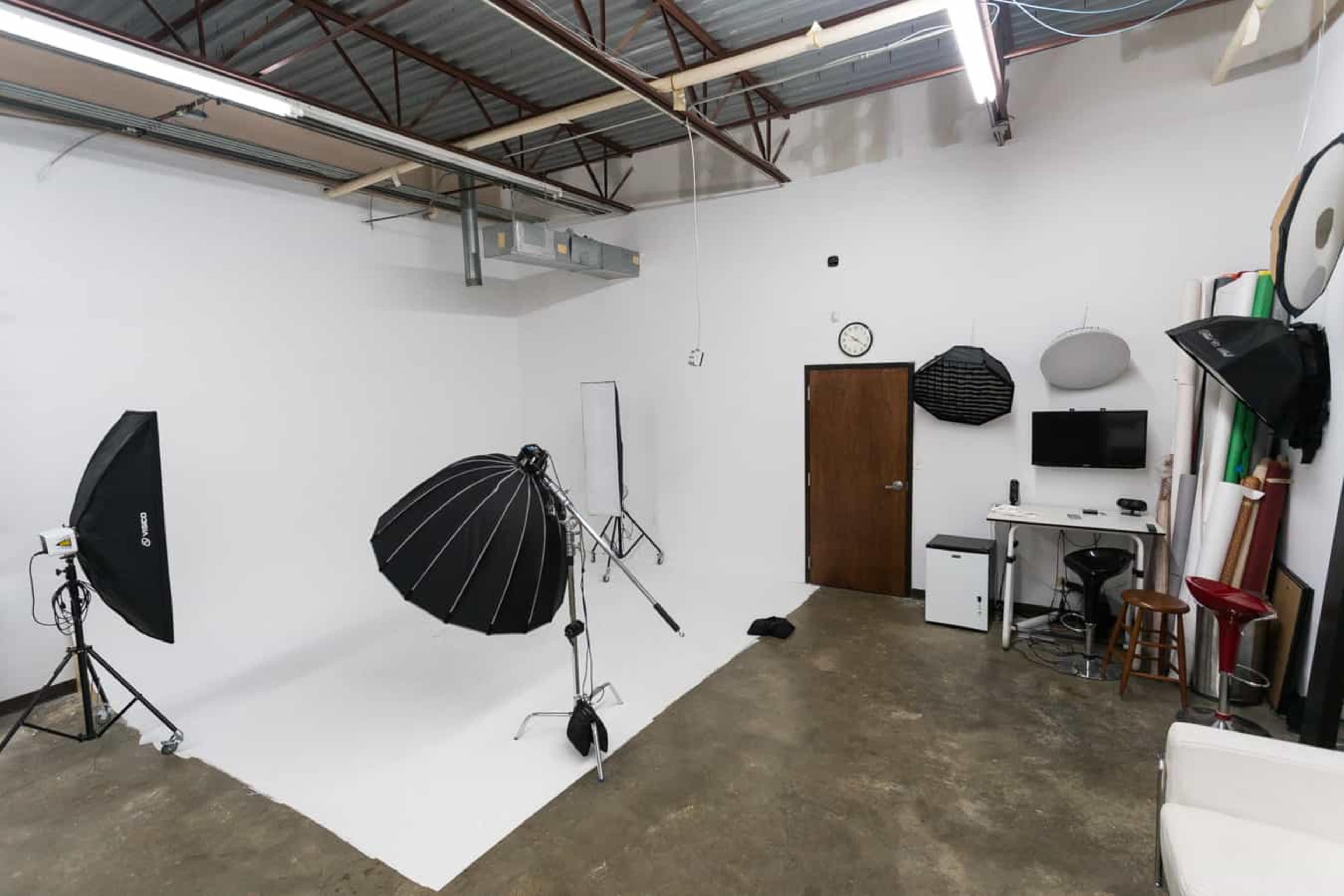 The image shows a photography studio with equipment such as softboxes, a backdrop, and a desk with a monitor.