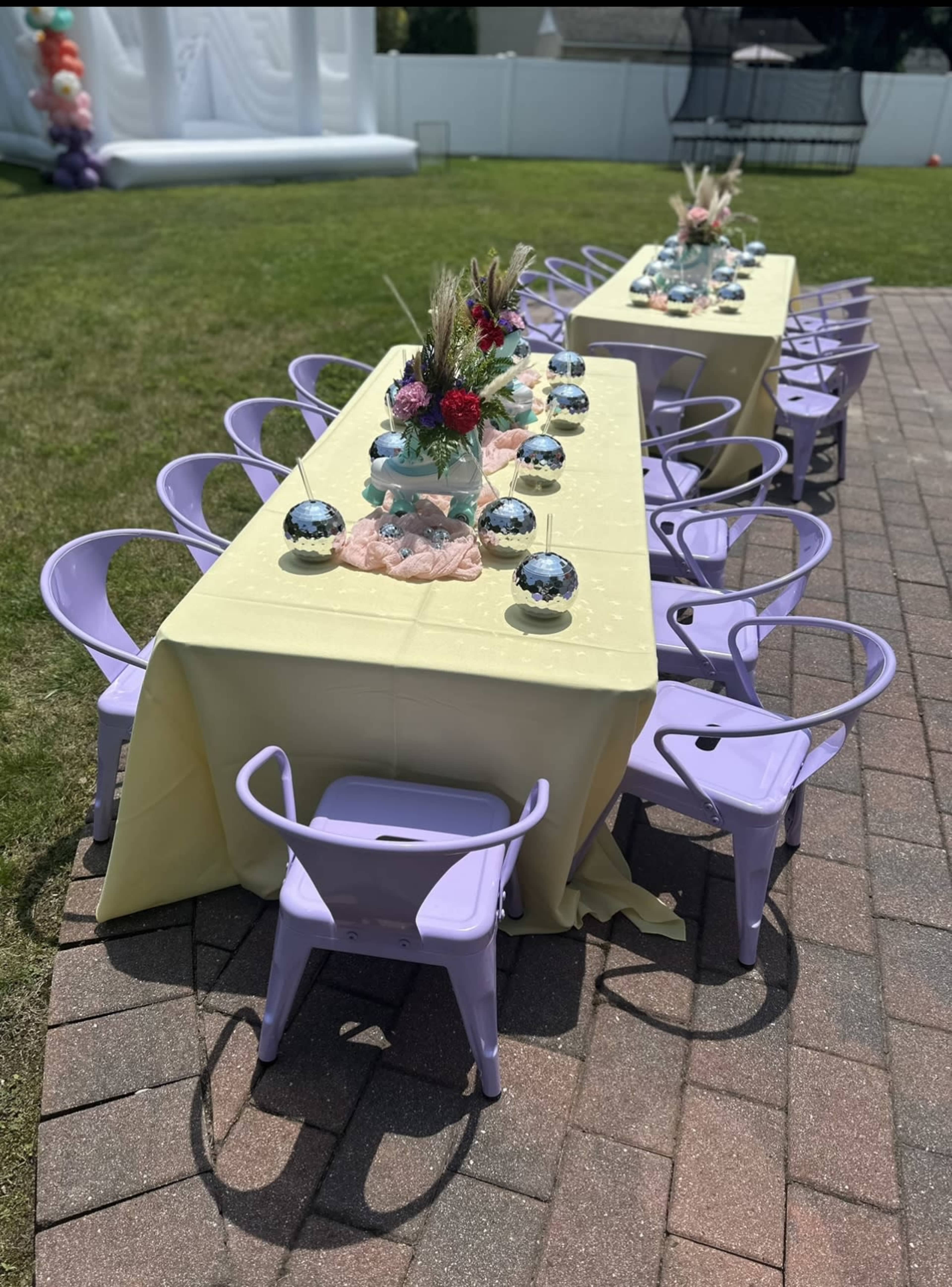 A long table set with decorative flowers and centerpieces is surrounded by purple chairs on a grassy area.