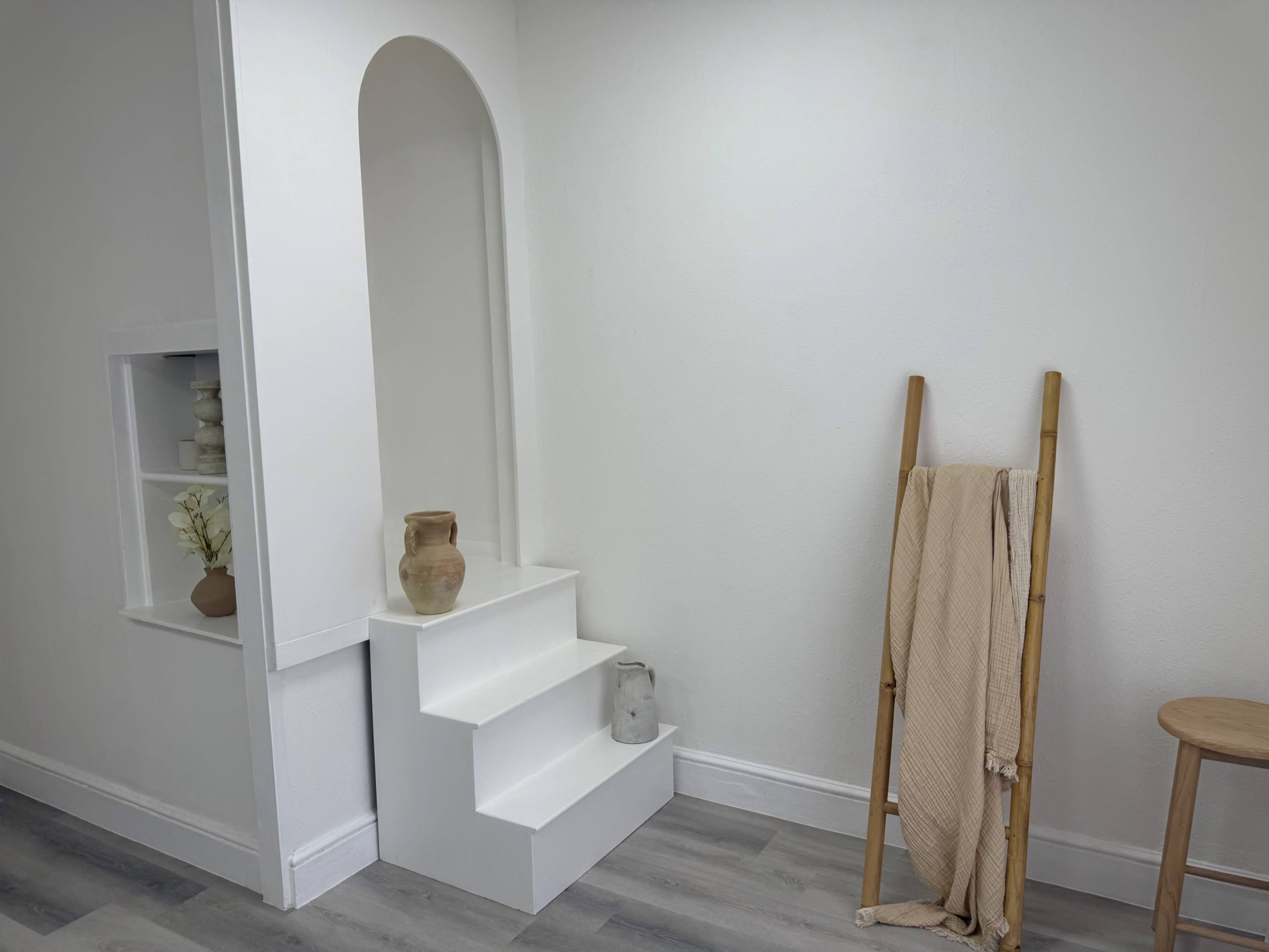 The image shows a minimalist interior featuring white steps leading to an archway, with a vase placed on the top step and a bamboo ladder holding a cream-colored blanket nearby.