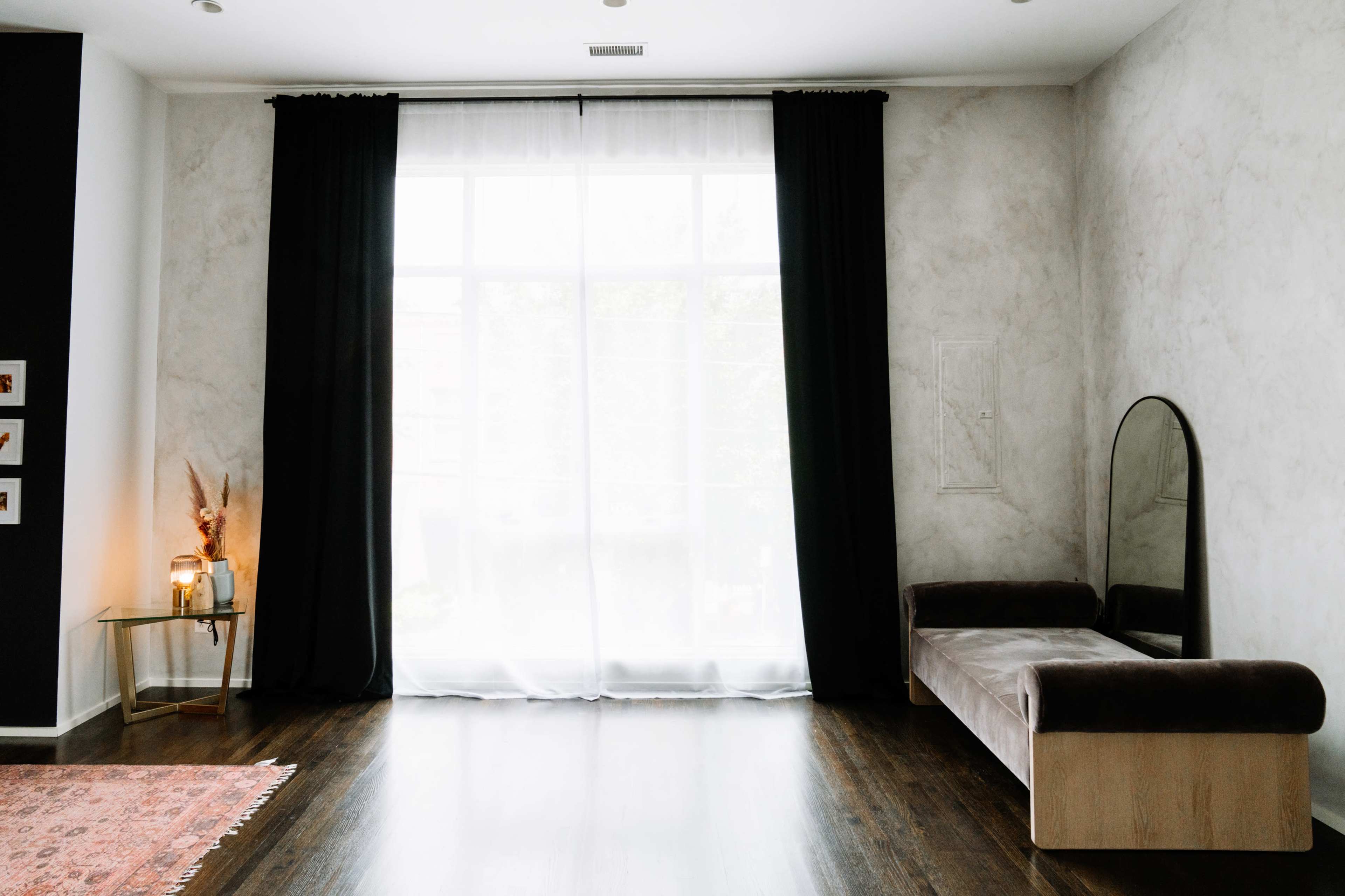 A minimalist interior features a curved bench, a round mirror, and a large window partially covered by black curtains.